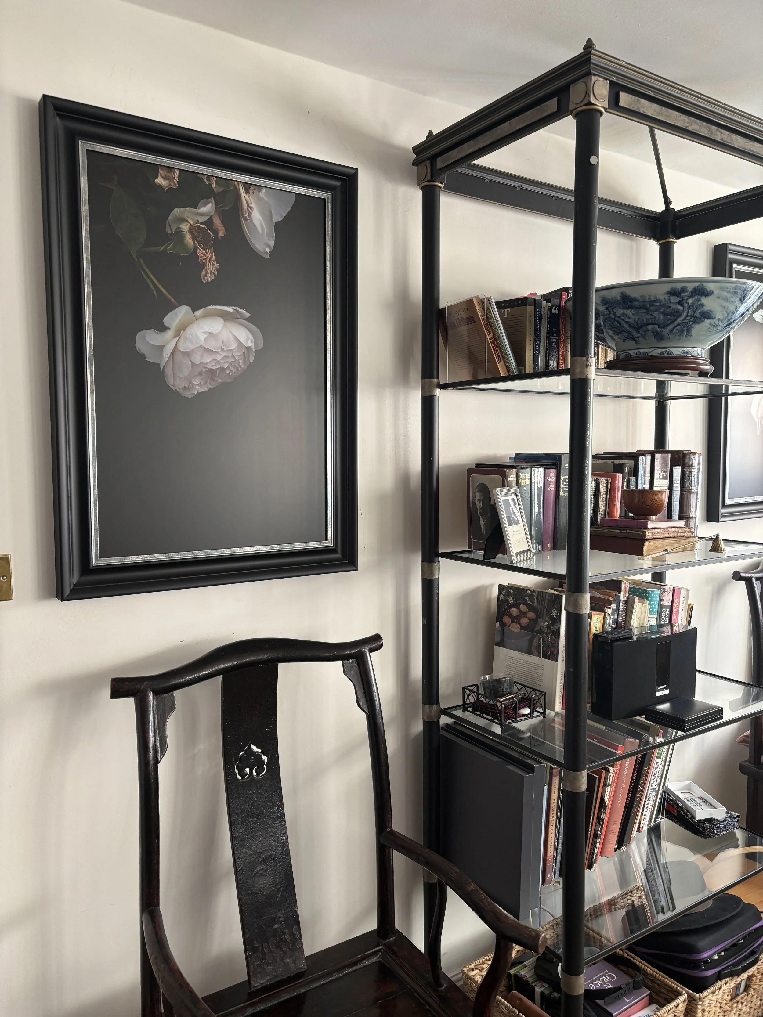 A dark wooden chair with a curved backrest in front of a cream-colored wall with a framed black background botanical print of a white flower with green leaves. A tall black metal and glass bookshelf with books, a bowl, and decorative items stands next to it.