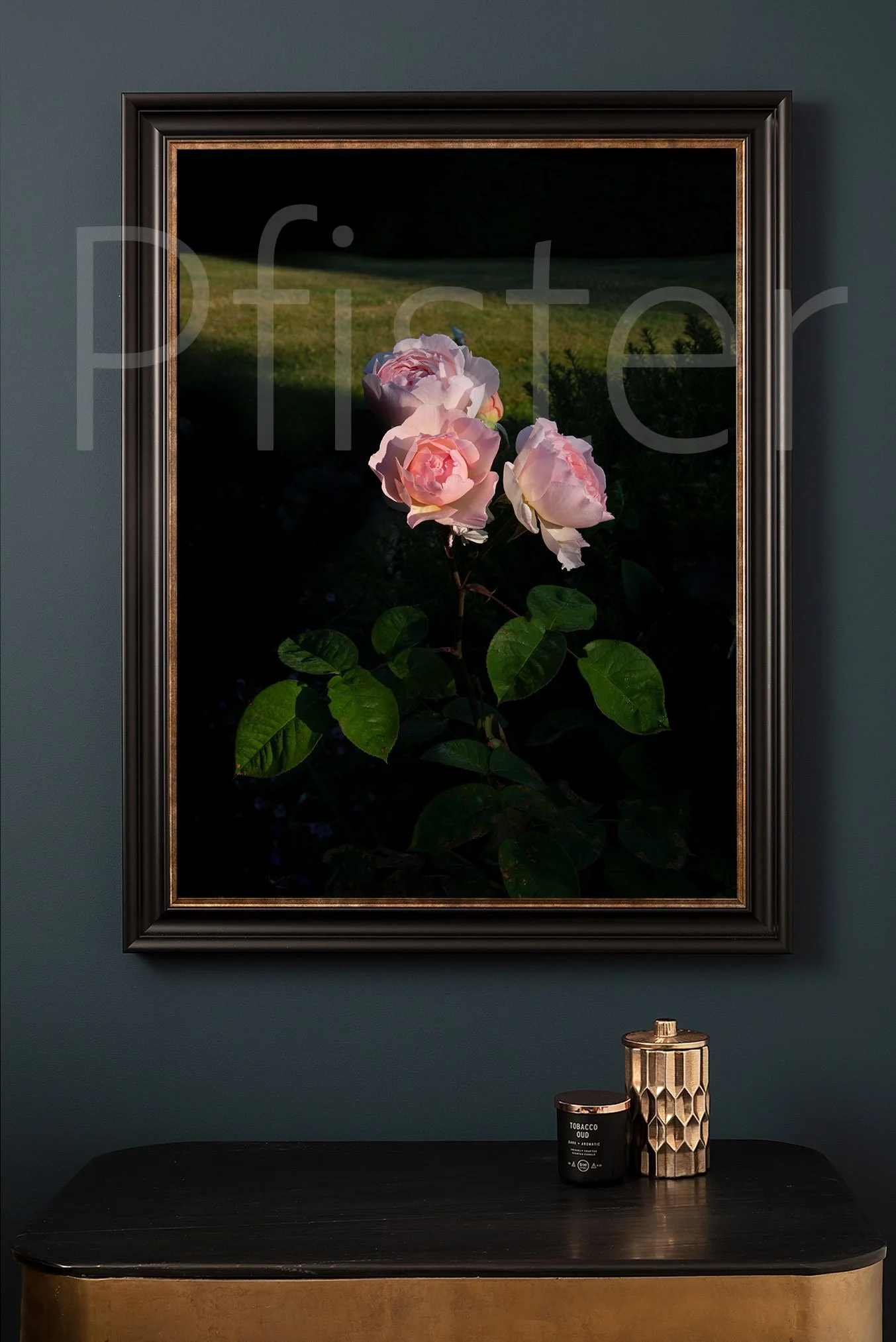 Framed art print of pink roses hanging on a dark wall, with a black and gold table below holding a black candle and a gold decorative container.