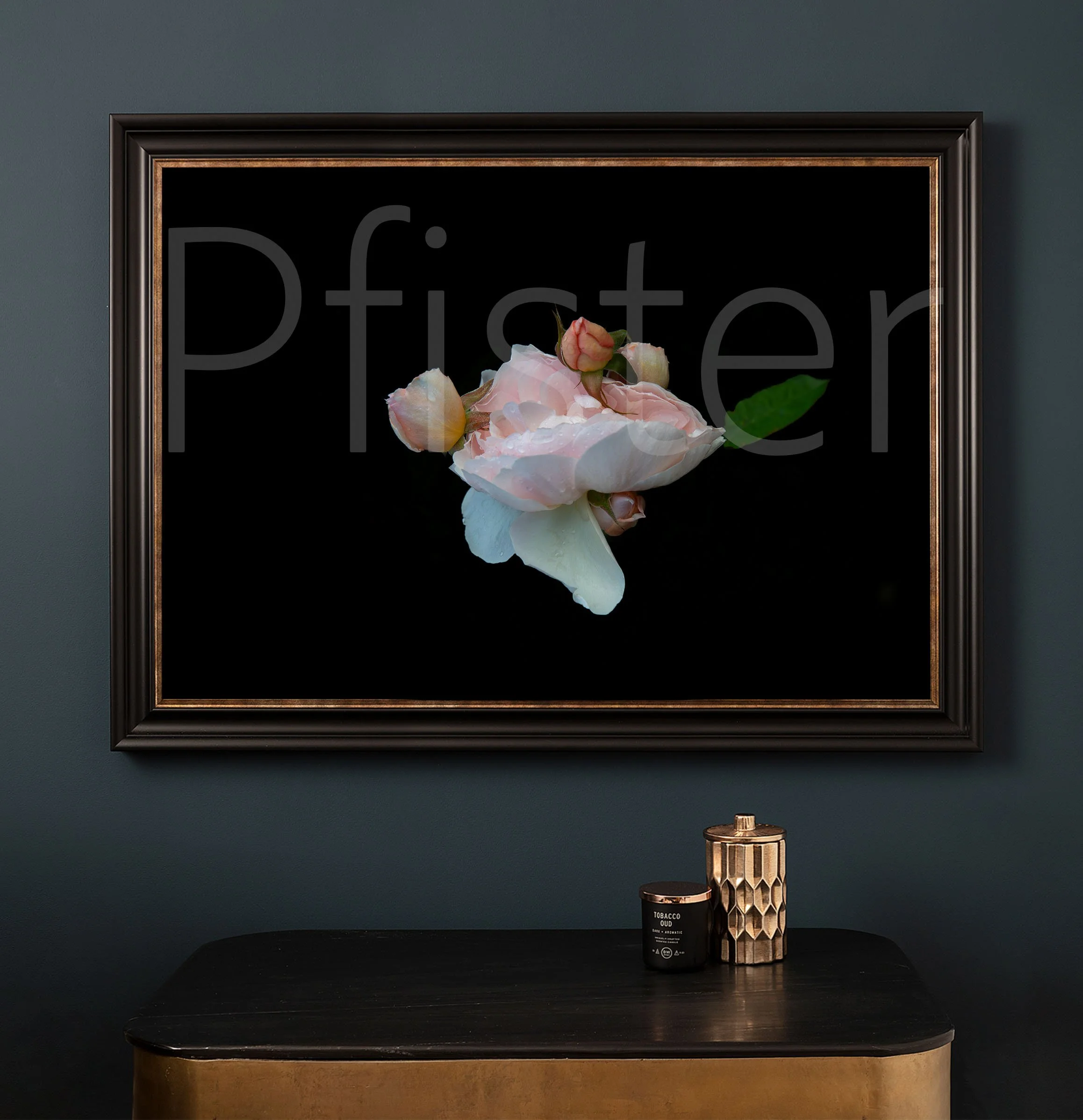 Framed artwork of a pink peony flower with buds on a black background, hanging on a dark green wall above a black and gold dresser with a black candle and a gold, textured container.
