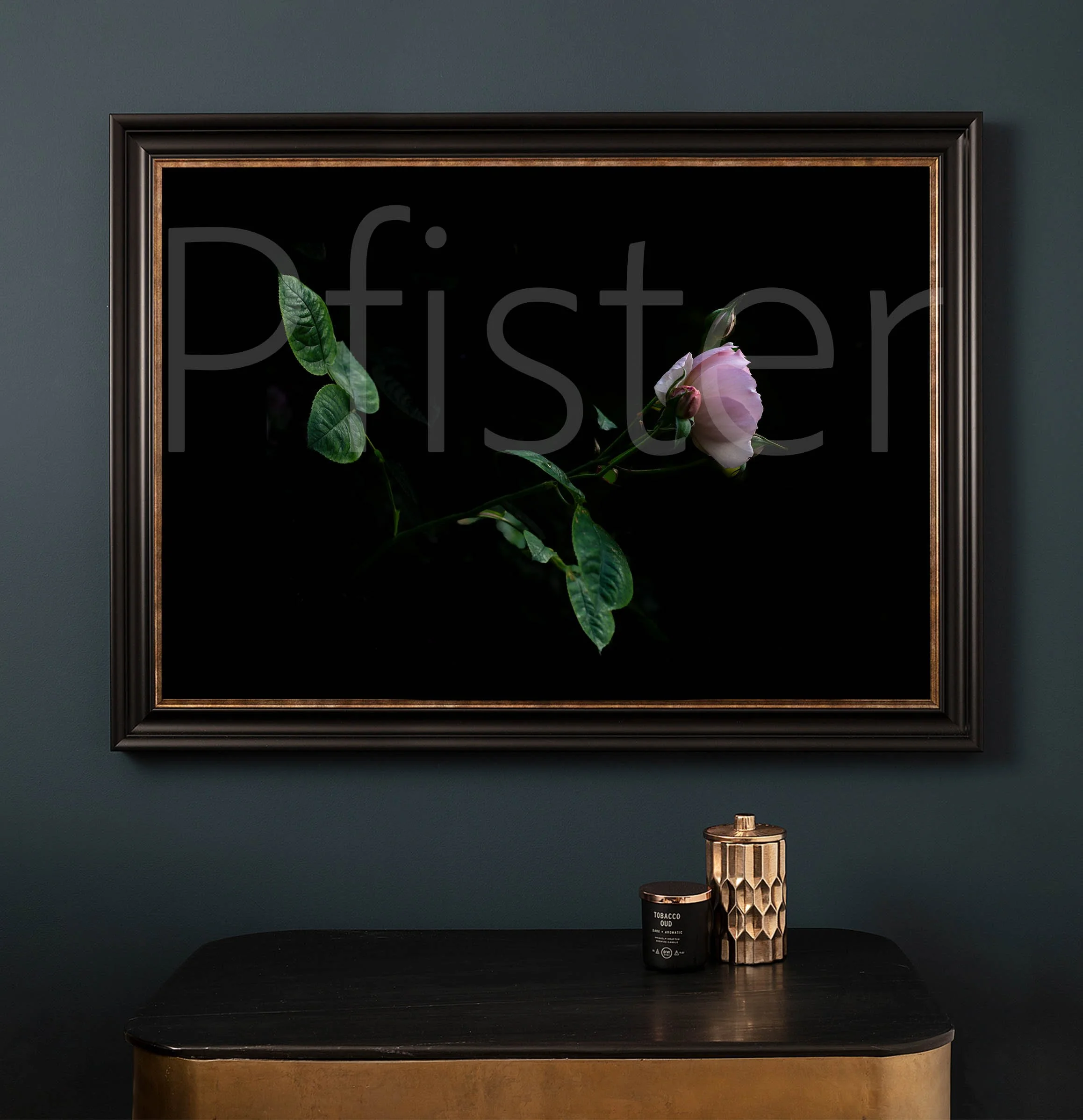 A black-framed painting of a pink rose with green leaves against a black background hangs on a dark wall. Beneath the painting, a black surface holds two decorative objects: a black candle with a label and a tall, textured gold vase.
