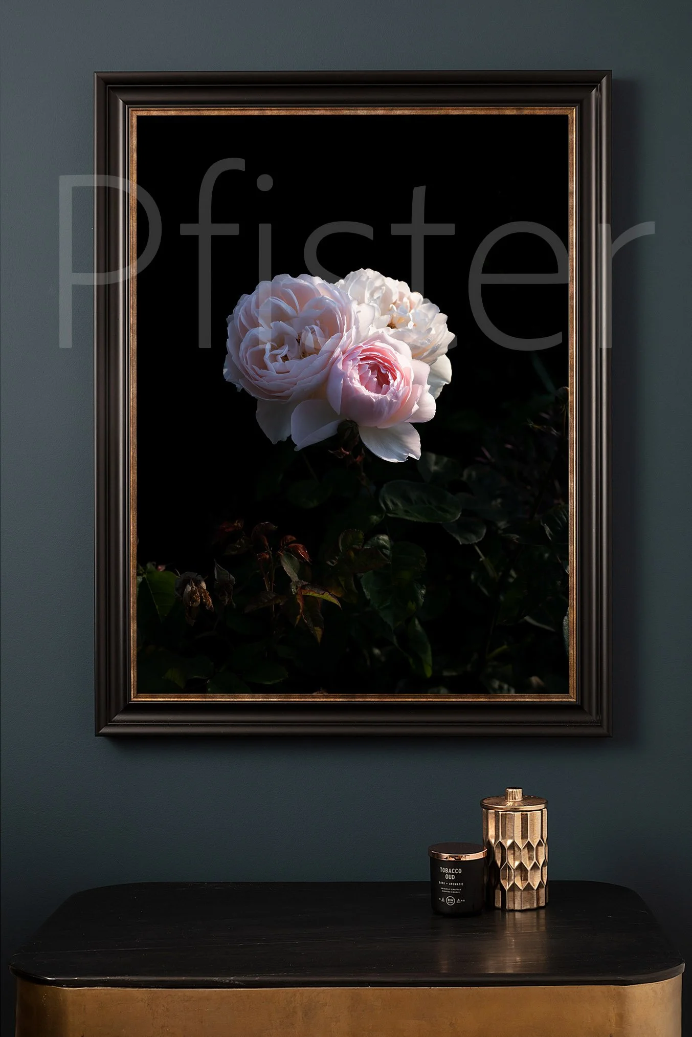 A framed floral photograph of white and pink peonies hanging on a dark green wall above a black table, with decorative objects including a candle and a geometric container.