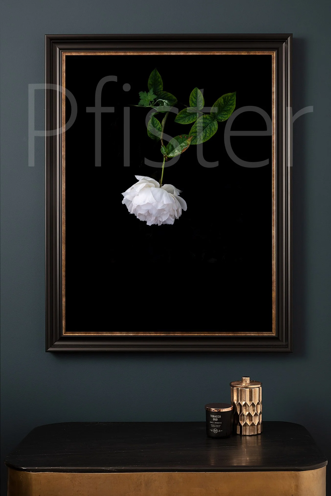 Wall art with a white flower and green leaves against a black background hanging above a black and gold table with two decorative objects, one black with gold accents and a warning label.