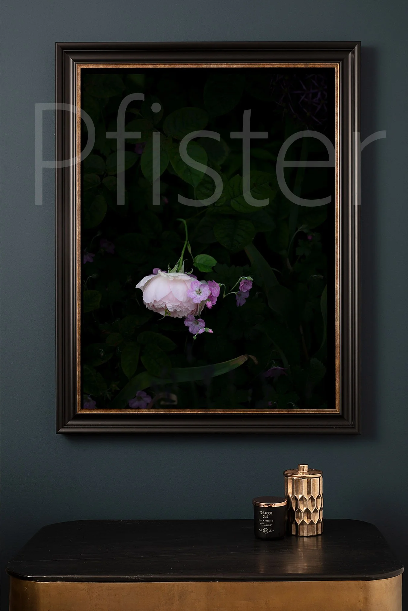 A framed dark floral picture hangs on a dark green wall. On a black and gold sideboard below it, there is a black candle with white text and a gold textured container.