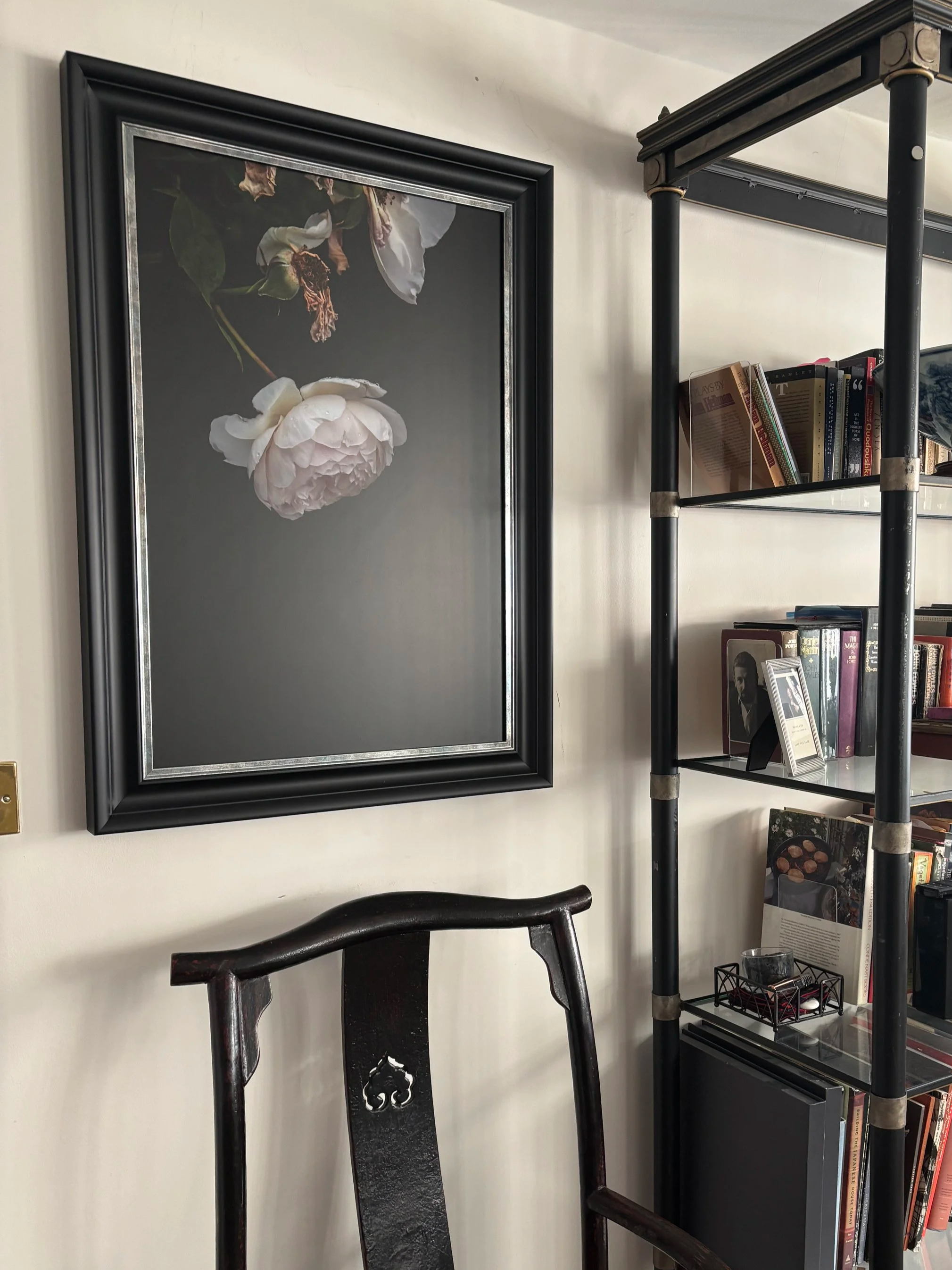 A dark wooden chair with a curved backrest in front of a cream-colored wall with a framed black background botanical print of a white flower with green leaves. A tall black metal and glass bookshelf with books, a bowl, and decorative items stands next to it.