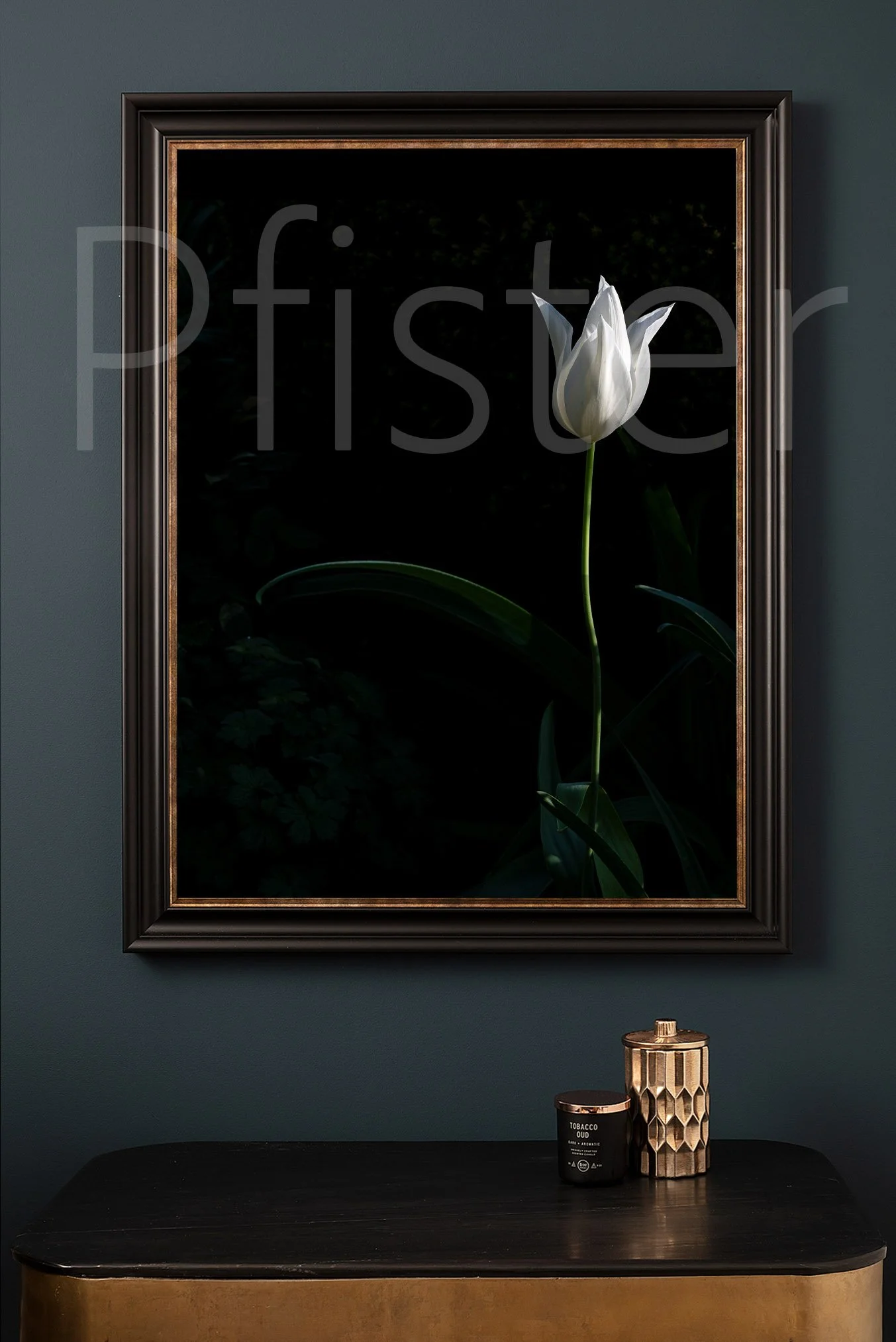 Framed black and gold floral artwork of a white tulip with green leaves on a dark background hanging above a black and gold side table, which holds a black candle and a gold textured container.