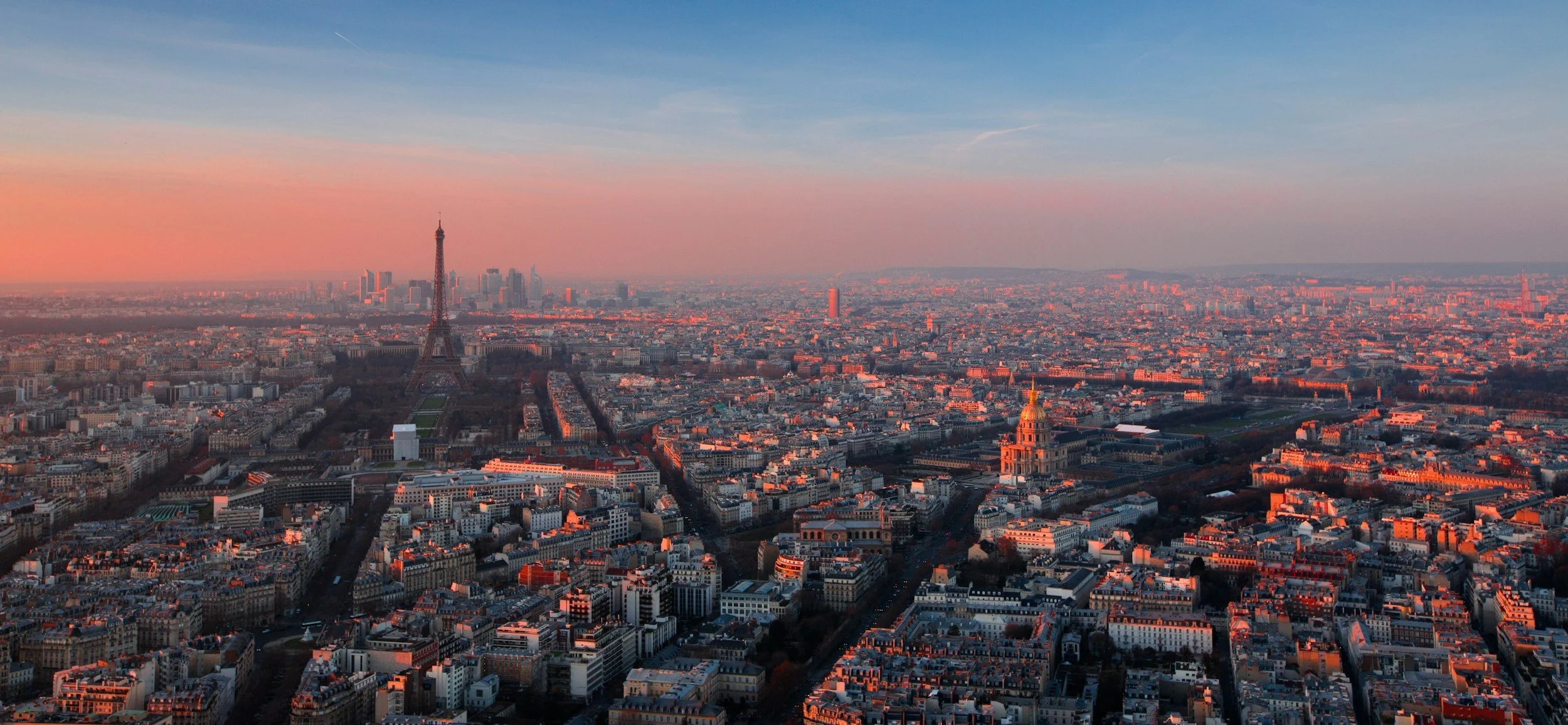 Vue aérienne de Paris au coucher du soleil, avec la Tour Eiffel au centre, entourée de bâtiments résidentiels et administratifs, sous un ciel aux teintes orange et bleues.