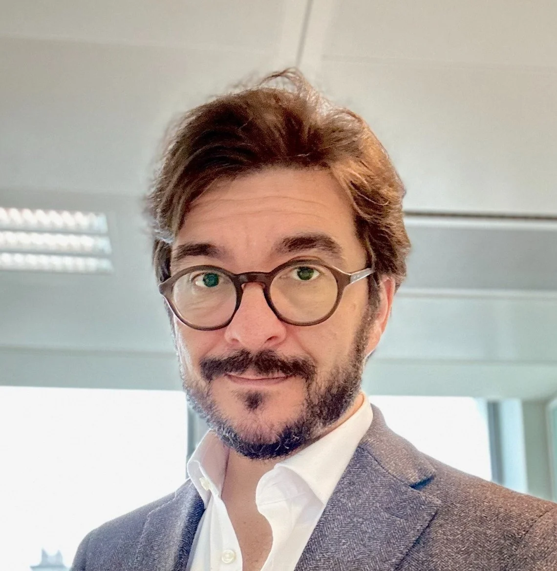 Portrait d'un homme avec des lunettes, moustache et barbe, portant un costume dans un environnement intérieur lumineux.