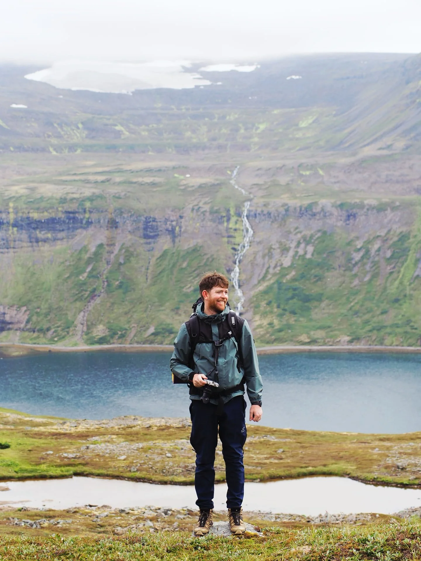 James Taylor, travel writer and photographer specialised in Iceland, standing on grassy terrain near a fjord with mountains and a waterfall in the background.