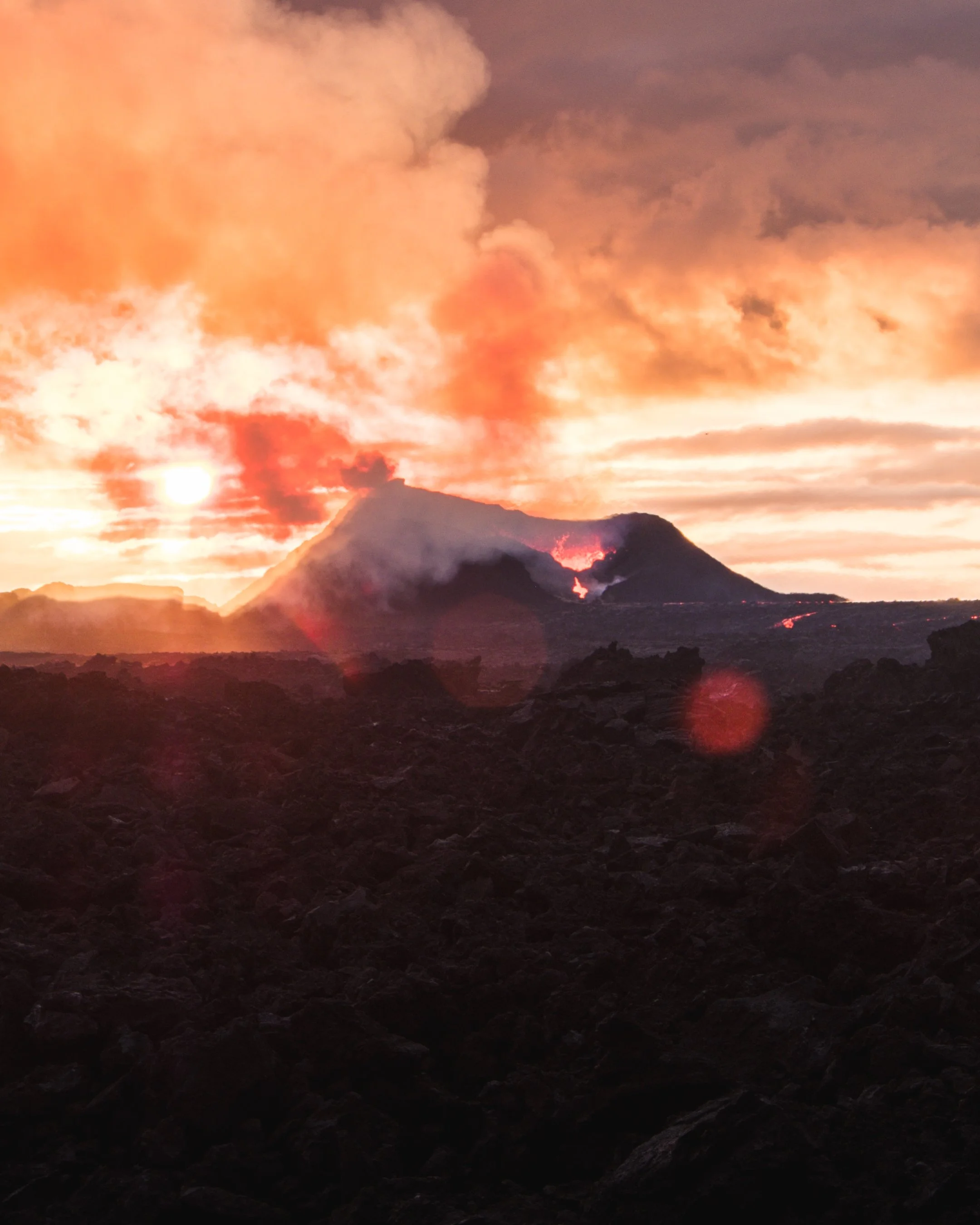 A volcanic eruption at sunset with smoke and lava, dark rocky foreground, vibrant orange and pink clouds in the sky.