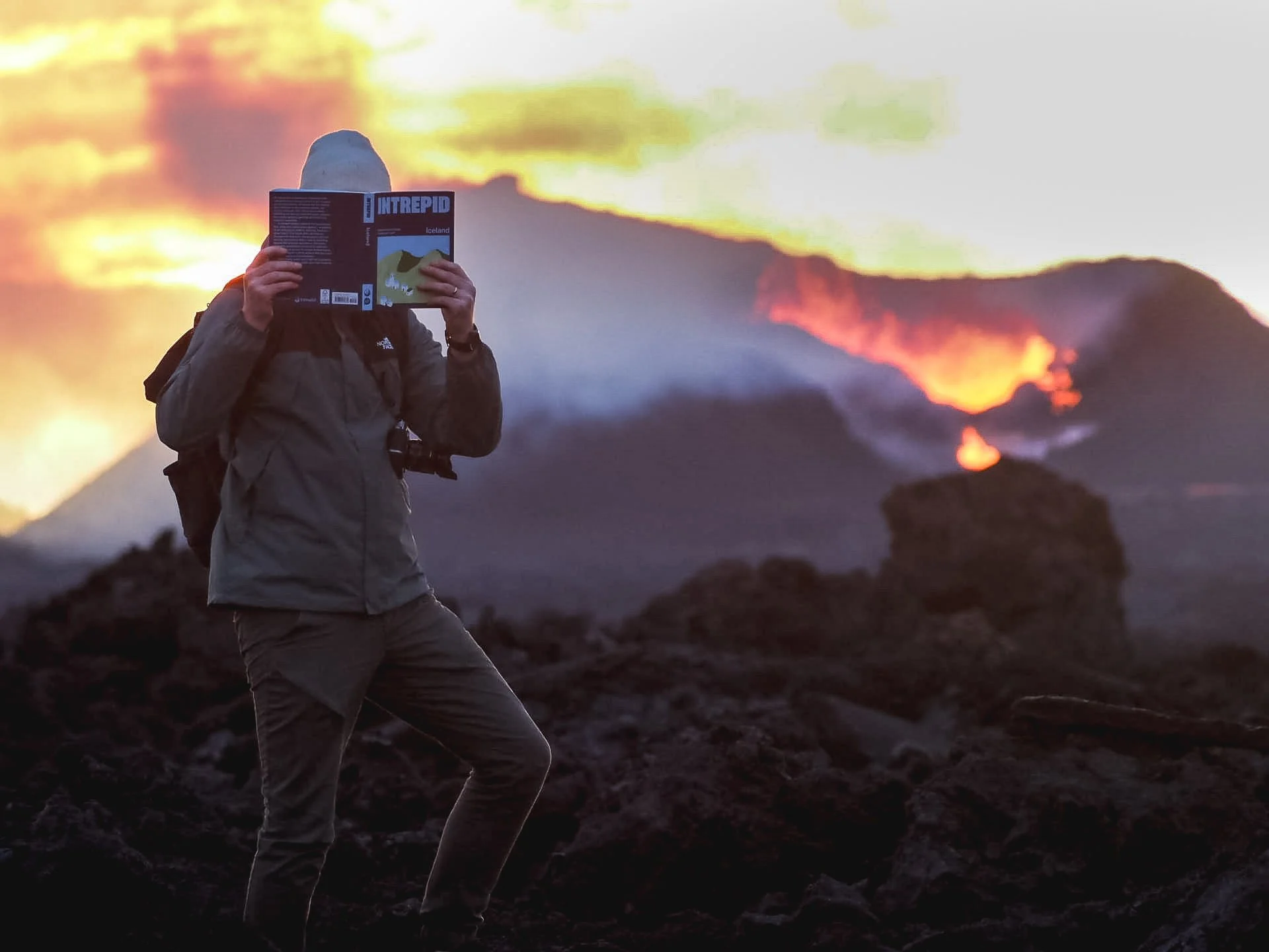 James Taylor, travel writer and photographer specialised in Iceland, standing on volcanic terrain, reading the Intrepid Guide to Iceland, with erupting volcano in background during sunset.