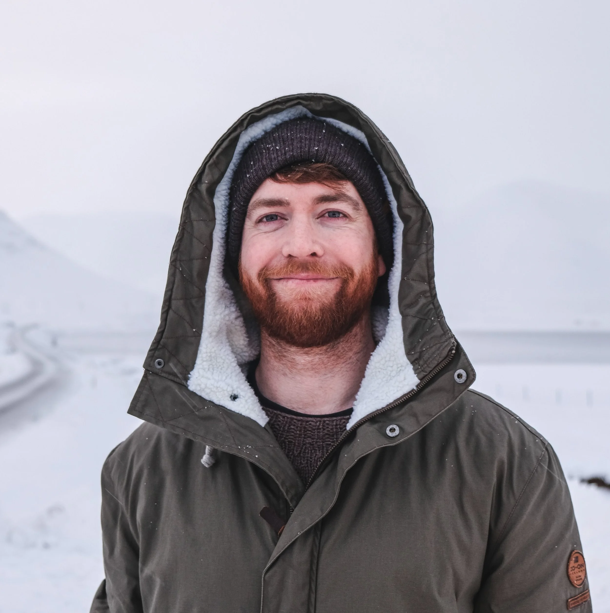 A man with a beard and mustache wearing a winter coat and a beanie, smiling outdoors in a snowy landscape.