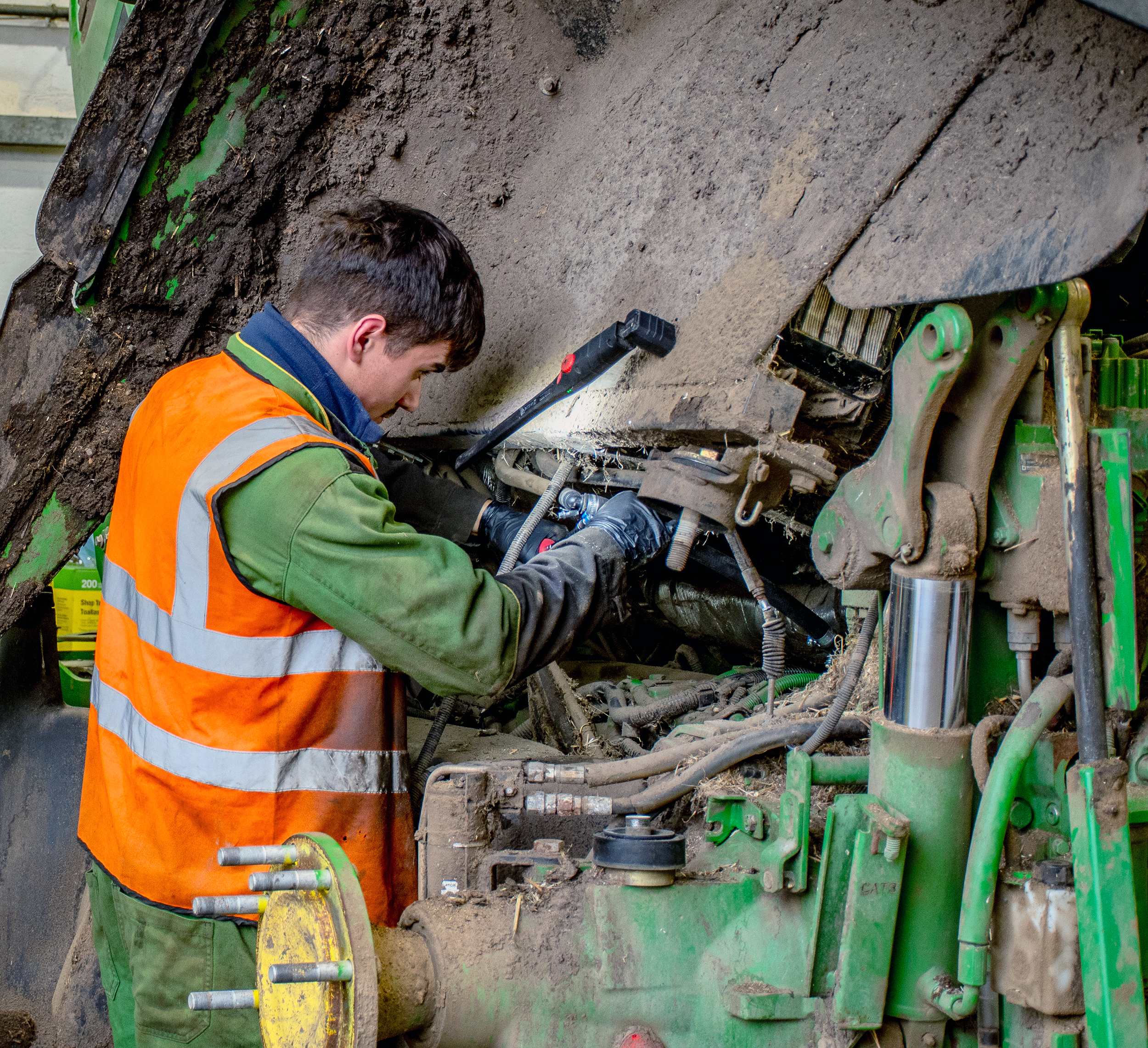 A worker wearing a high-visibility orange vest and green jacket repairs machinery