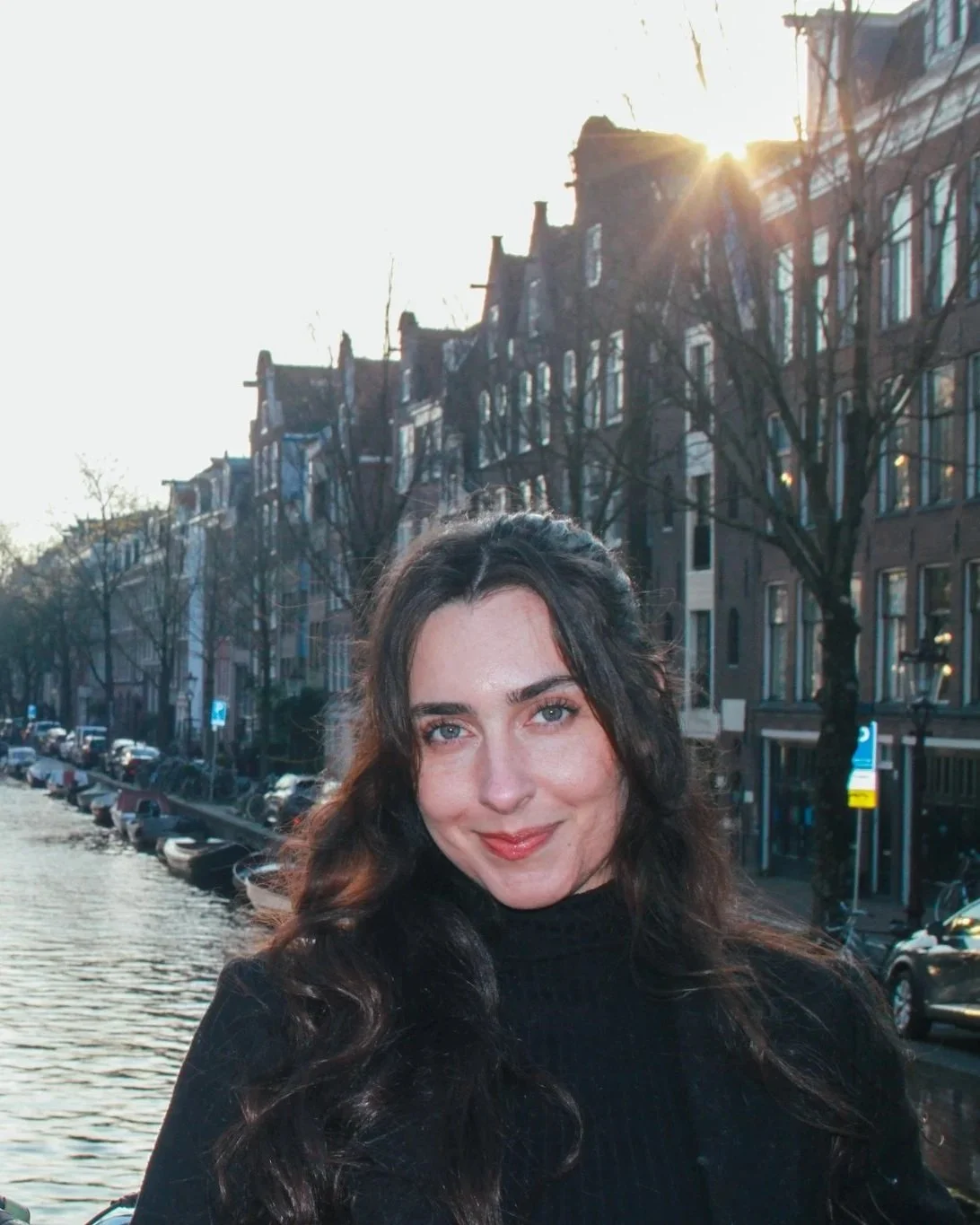 A woman with long dark hair and blue eyes smiling outdoors near a canal, with buildings and a tree-lined street in the background, and sunlight shining through the buildings.