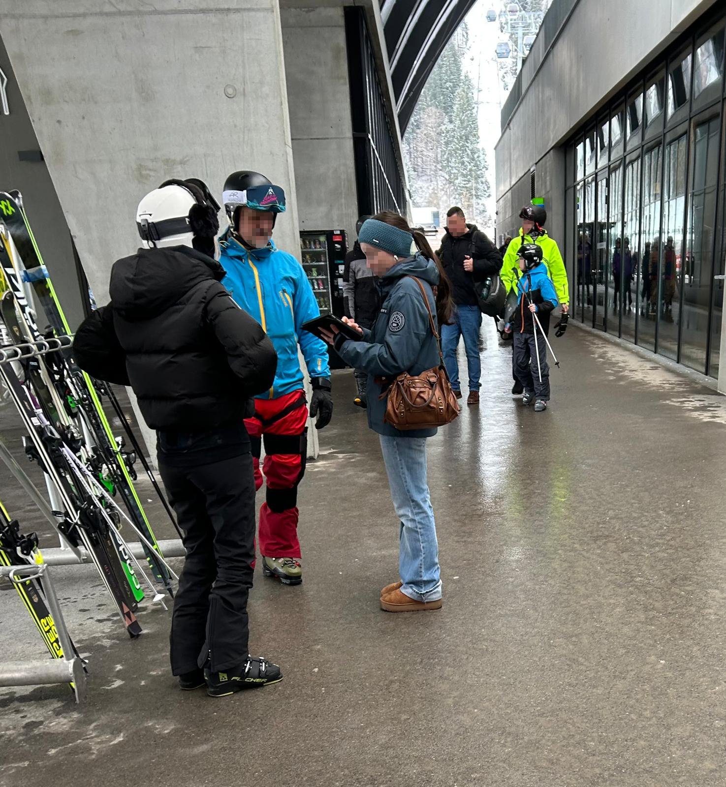 Eine Befragerin mit Tablet interviewt zwei Tourist:innen unter dem Vordach einer Seilbahntalstation. Einer der Tourist:innen trägt eine blaue Jacke mit roter Hose, die andere ist ganz in Schwarz gekleidet. Beide tragen einen Skihelm.