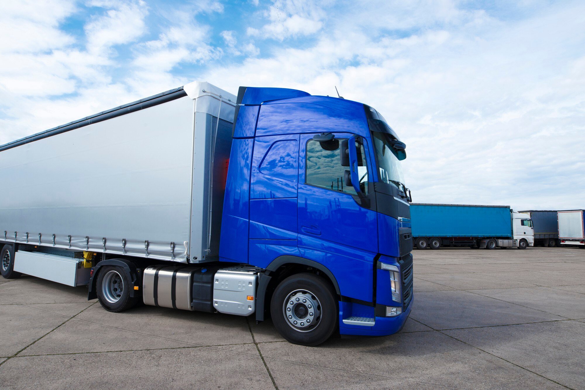 Blue semi-truck parked on a concrete lot with additional trucks in the background, under a partly cloudy sky.