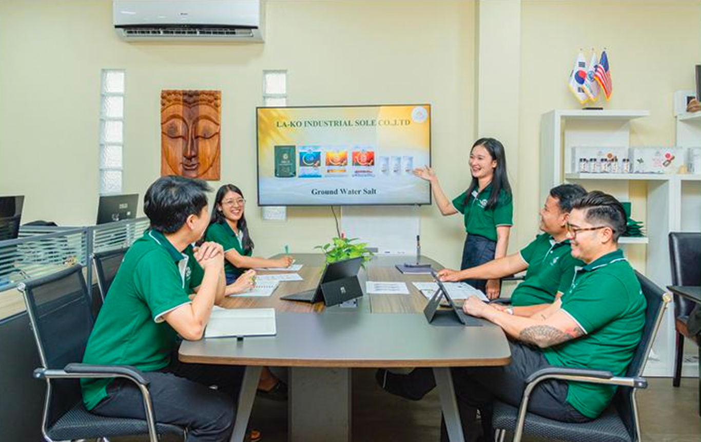 People attending a presentation in an office conference room. A woman stands pointing to a large screen displaying a presentation. Five people seated at the table are looking at her and smiling, with laptops and notebooks in front of them.