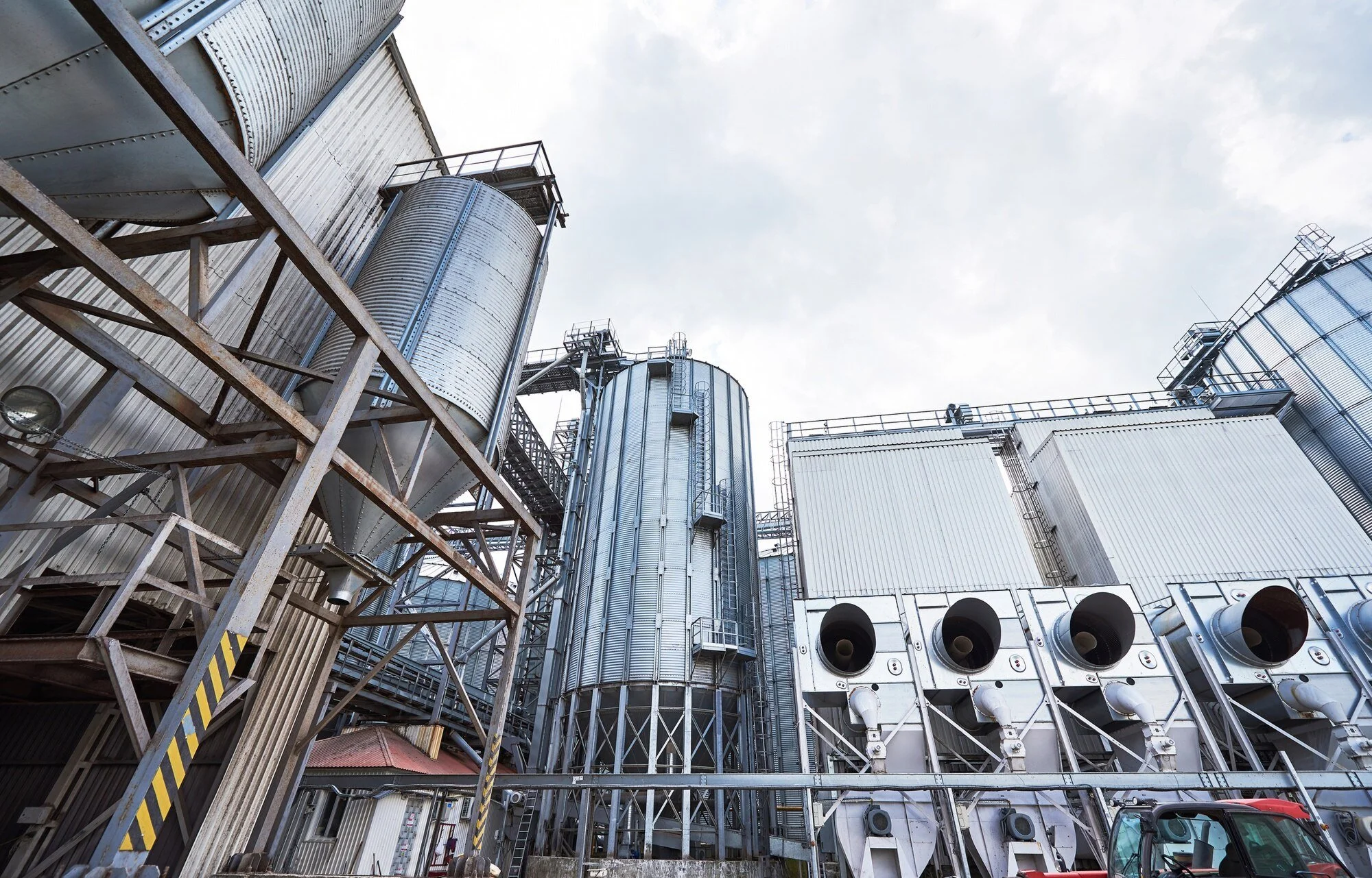 Industrial facility with large metal silos, ducts, and structures under a cloudy sky.