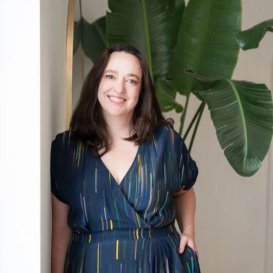 A woman with long brown hair wearing a dark blue dress with colorful vertical stripes, standing in front of a large green plant indoors.