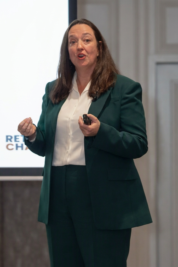 A woman with long brown hair wearing a green blazer and white shirt is giving a presentation, standing in front of a screen.