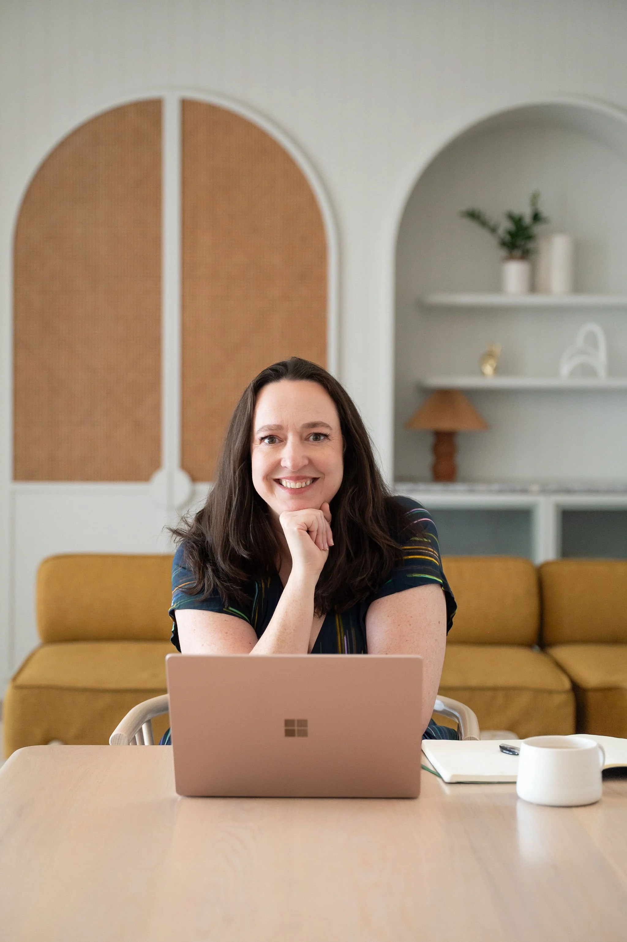 A woman with dark brown hair, wearing a dark top with colorful stripes, sits at a light-colored table in a room with a yellow couch and white shelves. She is smiling and resting her chin on her right hand while working on a pink laptop. On the table are a white mug, a small object, and a closed notebook.