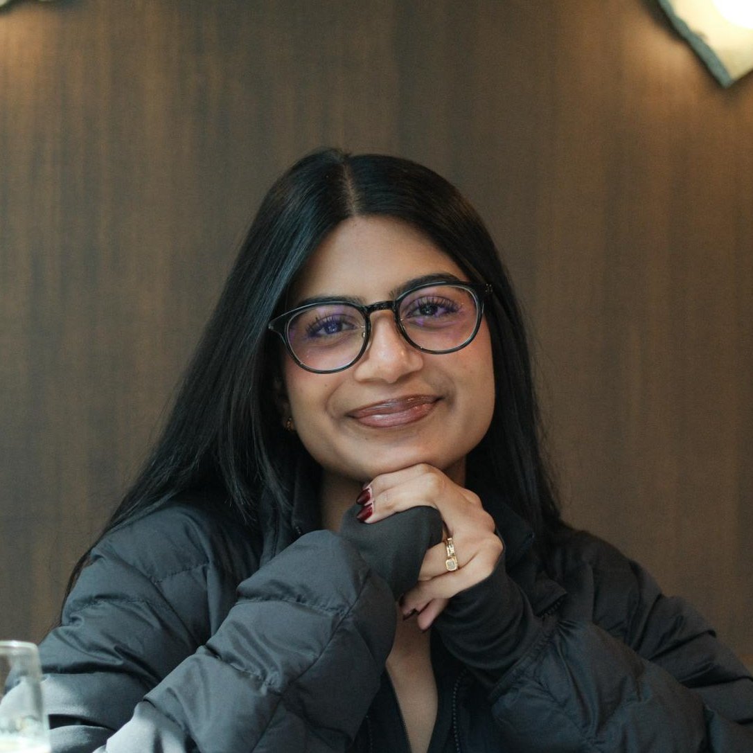 Clinical psychologist with long dark hair and glasses smiling with her chin resting on her hands, photographed against a dark brown wall.