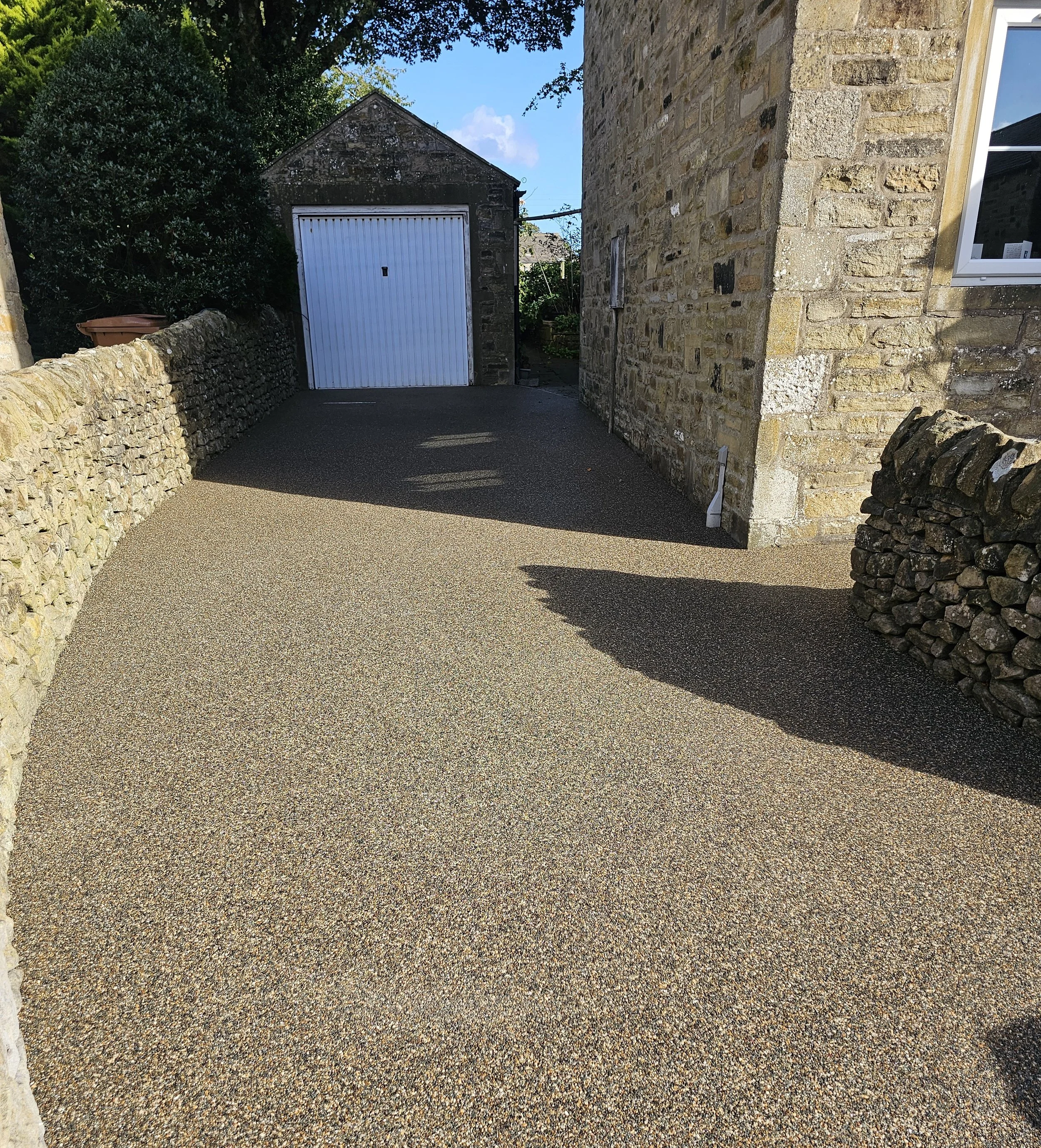 An outdoor scene showing a stone driveway leading to a garage with a white door, surrounded by stone walls and greenery, under a partly cloudy sky.