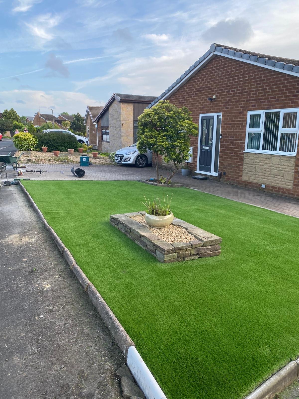 A front yard with a new green artificial grass lawn, a brick flower bed with a white pot, and a brick house with a blue front door and white window frames.