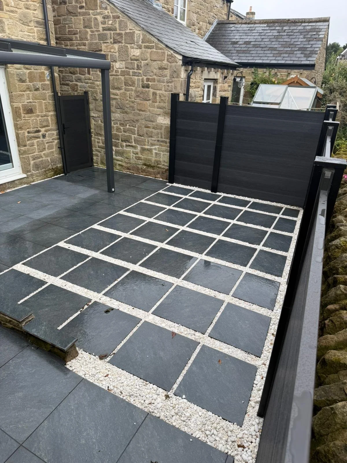 A small outdoor area with dark slate tiles and a grid of lighter, smaller stones in a square pattern. A black fence separates the space from neighboring properties, and stone walls and stone buildings are visible in the background.