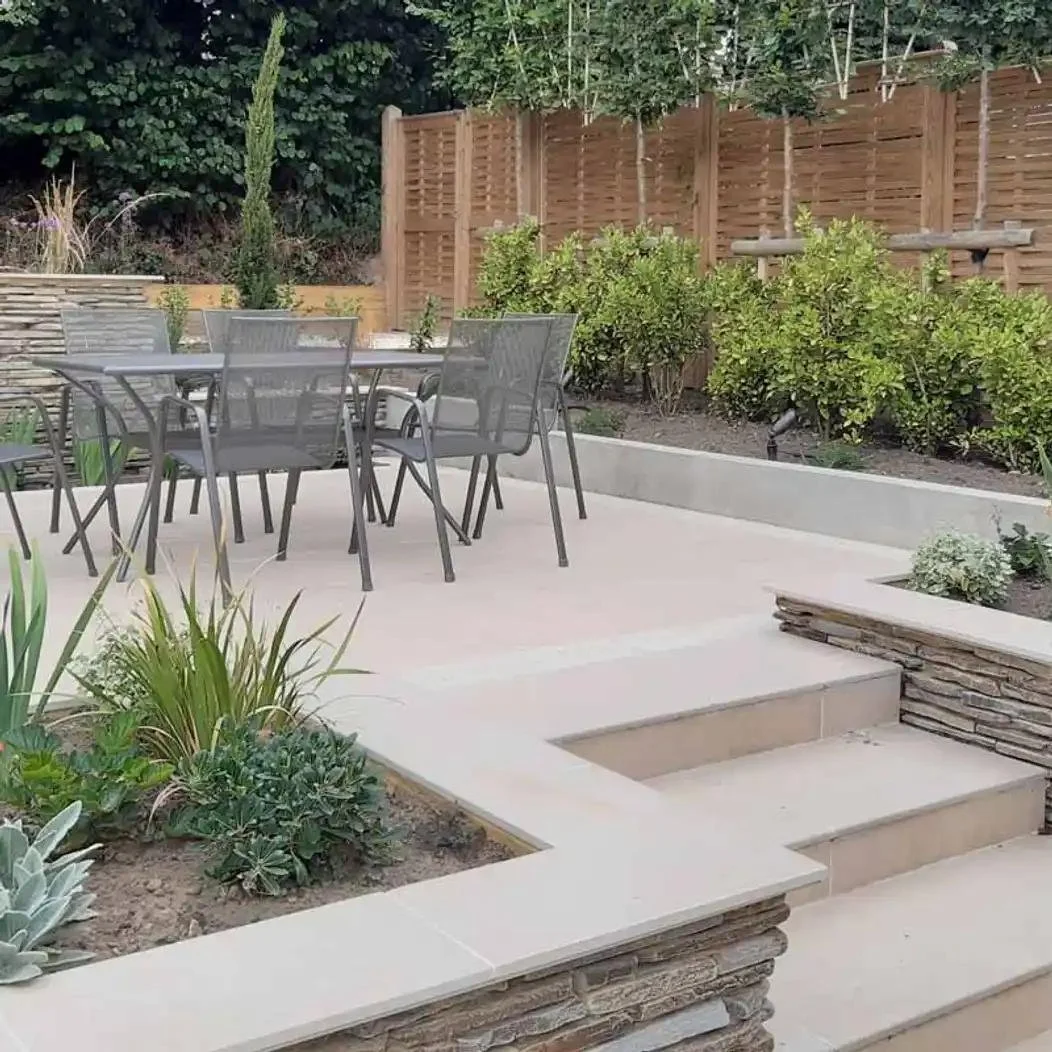 A backyard patio with outdoor dining table and chairs, surrounded by landscaped garden beds and a wooden privacy fence.