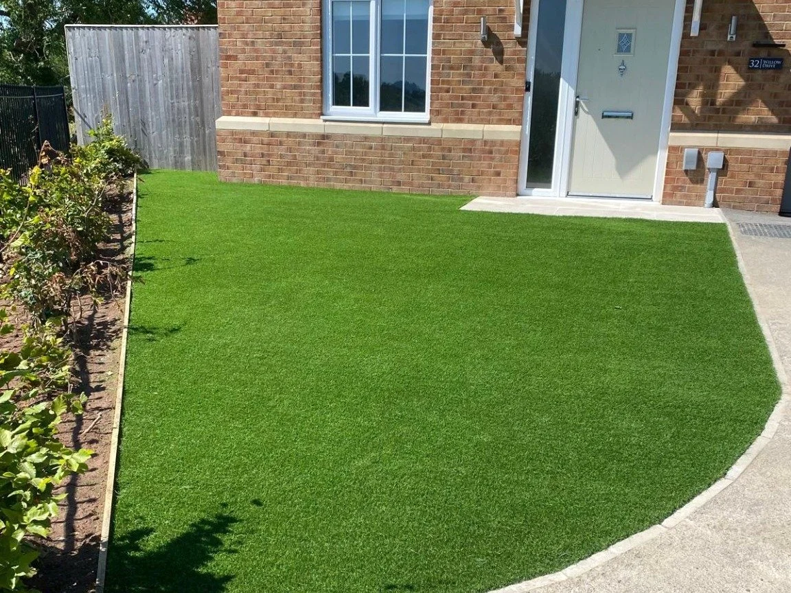 Artificial grass lawn next to a brick house with a window and a white door, bordered by a concrete pathway and a garden bed with plants.