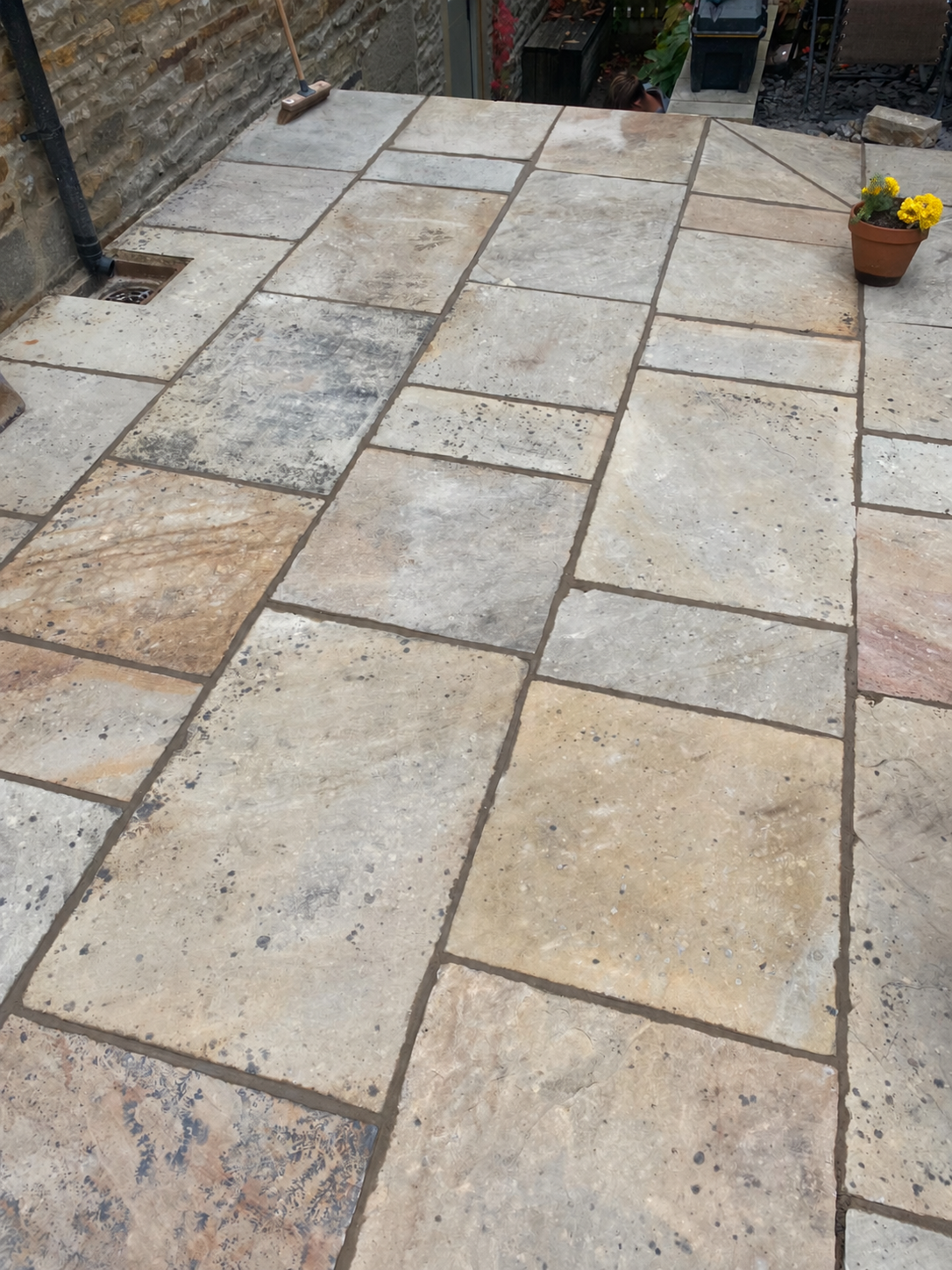 Close-up view of a stone patio with rectangular and square tiles in various shades of beige, gray, and cream, with some tiles featuring speckled and textured patterns. There is a potted yellow flowering plant on the right side of the image.