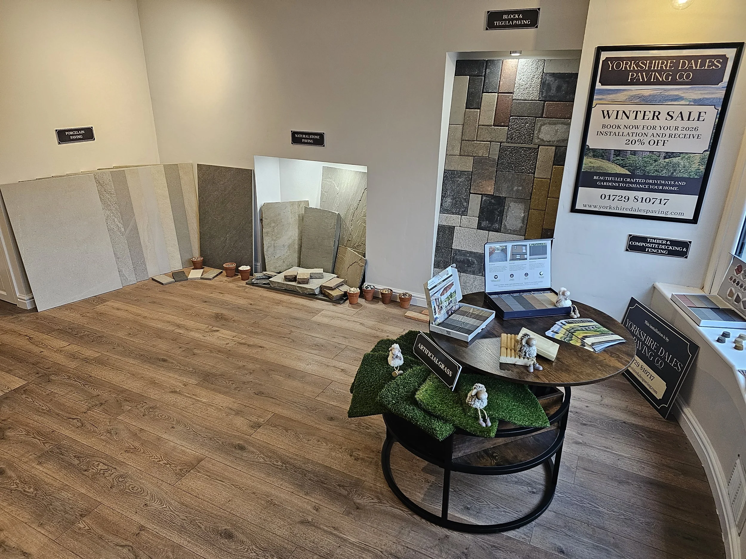 Showroom display of various stone and paving samples with a table holding brochures and decorative items, and promotional signs for paving services.