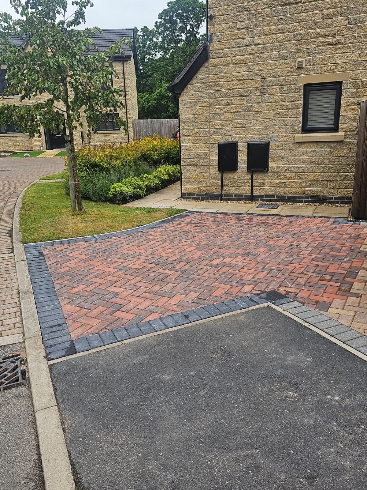 View of a newly paved driveway with red and gray bricks in front of a beige brick house, with a tree and landscaped garden on the side under an overcast sky.