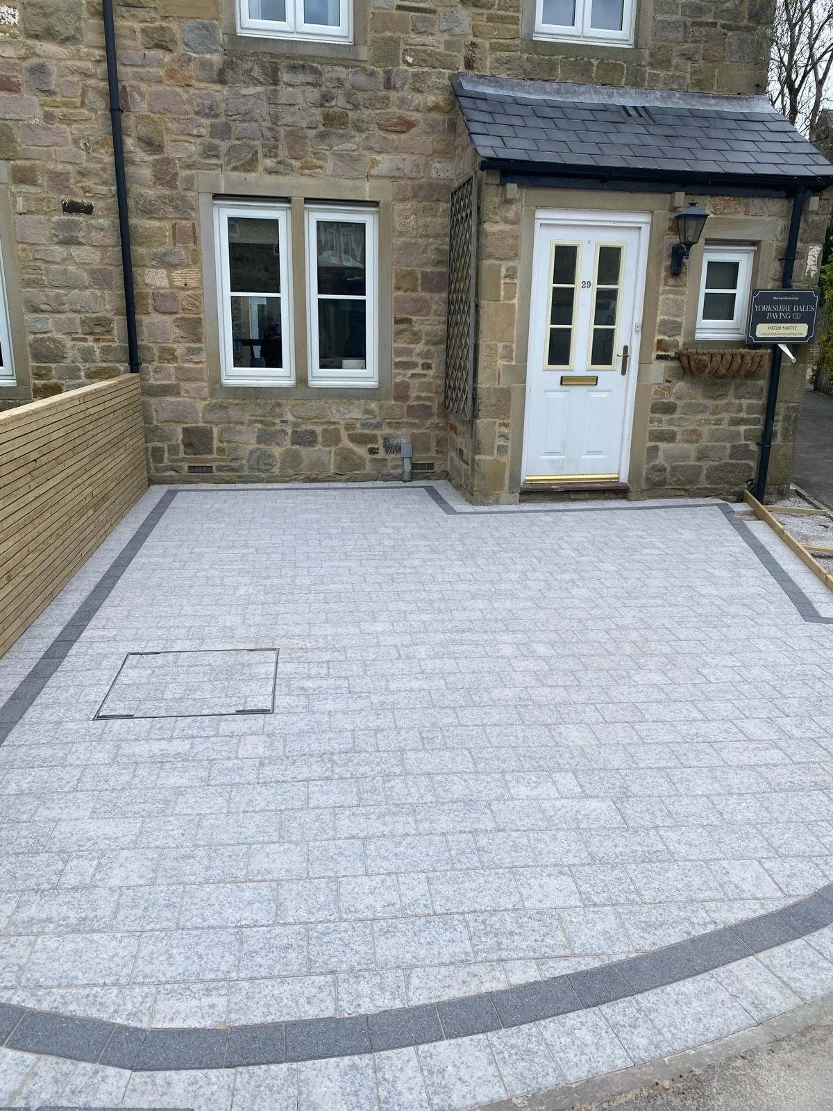 Front view of a small paved patio area outside a stone house with a white door, black lantern, two small windows, a wooden fence on the left, and a sign for Yorkshire Dales Paving Co.