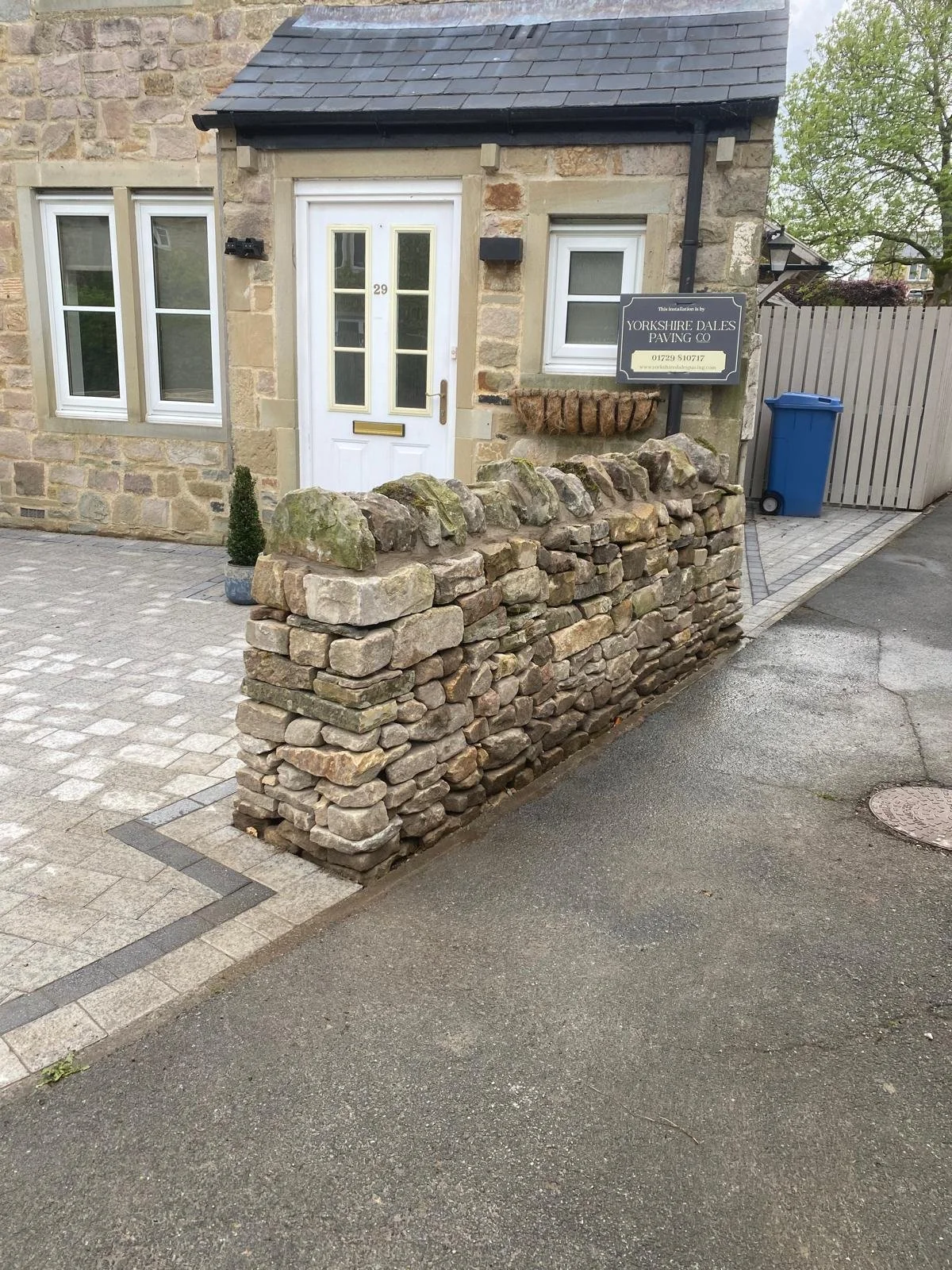 A stone wall encloses the front of a small stone house with a white front door and two windows. A sign on the wall indicates the installation is by Yorkshire Dales Paving Co. There is a small potted tree made of greenery next to the door. A gravel driveway and a paved sidewalk are in the foreground, with a blue trash bin near a wooden fence and a street lamp.