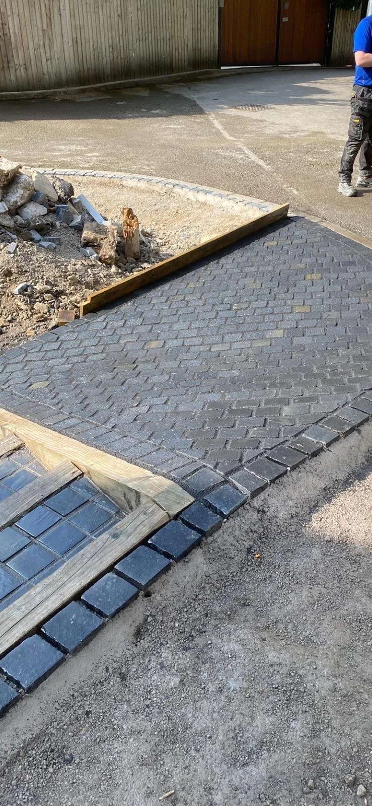 Landscape view of a driveway under construction, showing a freshly paved brick pathway with a curb border, adjacent to an unpaved dirt area, with a wooden board and construction debris nearby, and two workers in the background.