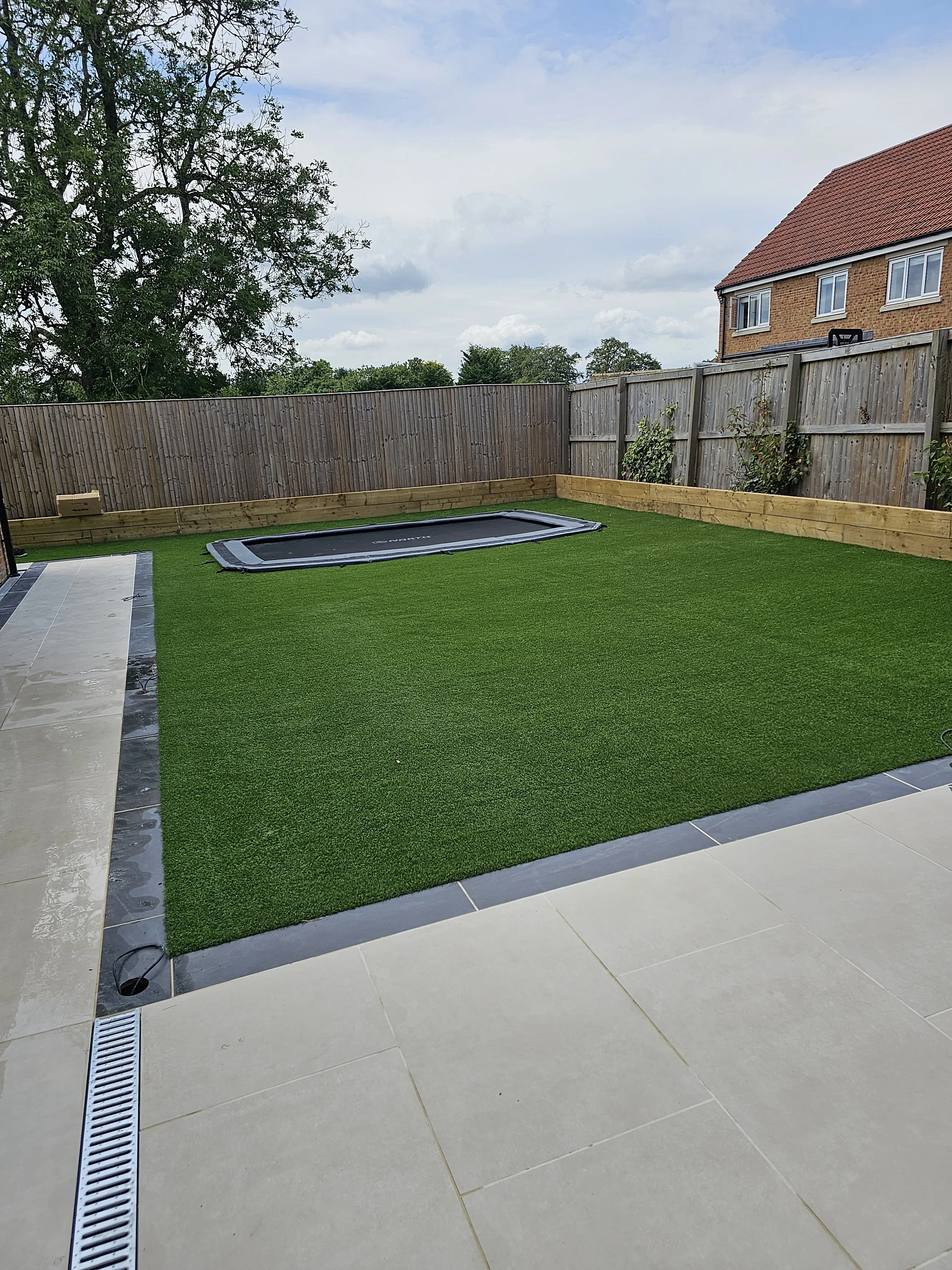 A backyard with a green artificial turf grass area, surrounded by a wooden fence, a trampoline on the turf, and a part of a tiled patio of premium porcelain in Ivory and Anthracite with a drainage grate and a water drain in the foreground.