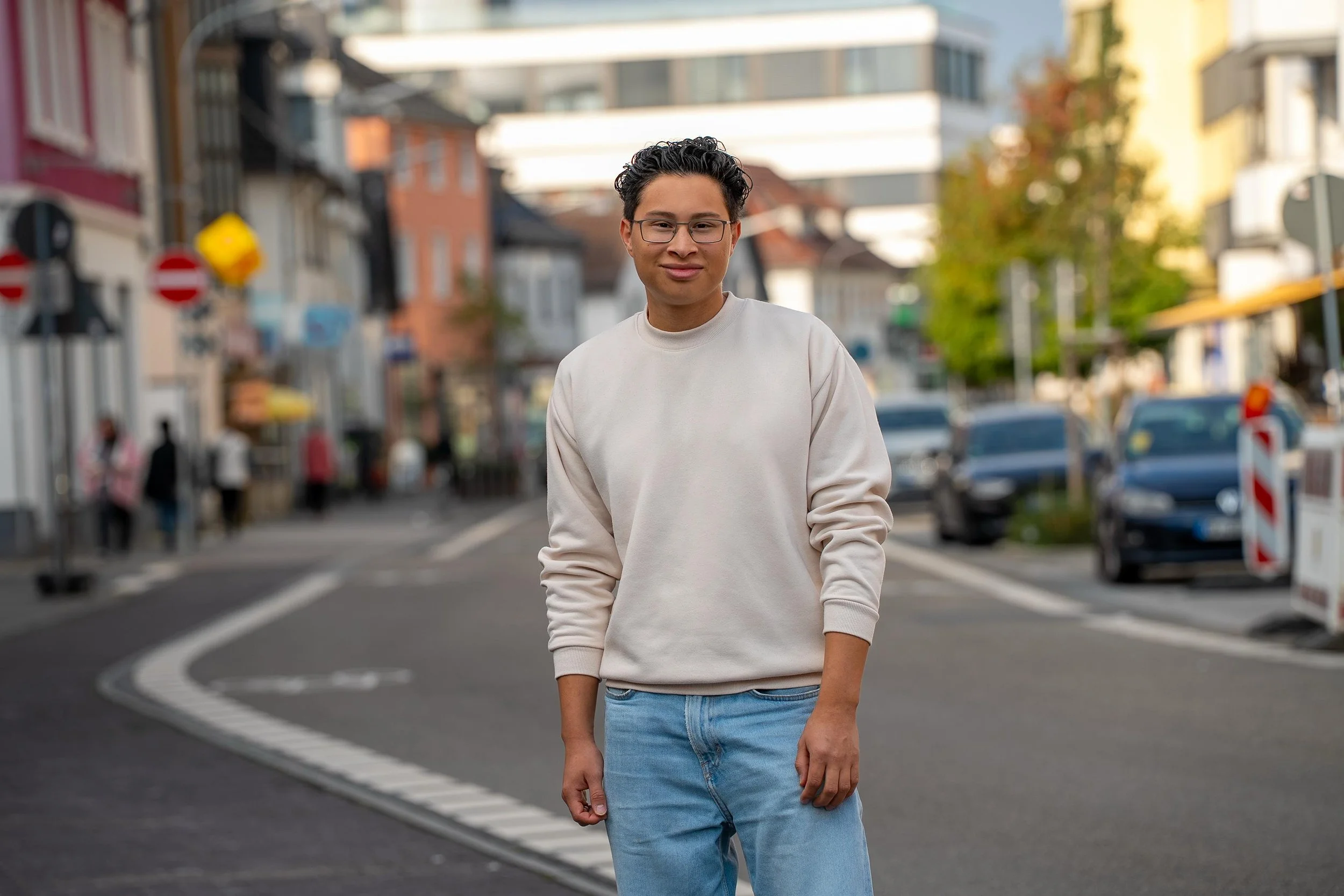 Junger Mann mit Brille in weißem Sweatshirt und Jeans steht auf einer Straße in einer Stadt, im Hintergrund sind Häuser und Autos sichtbar.