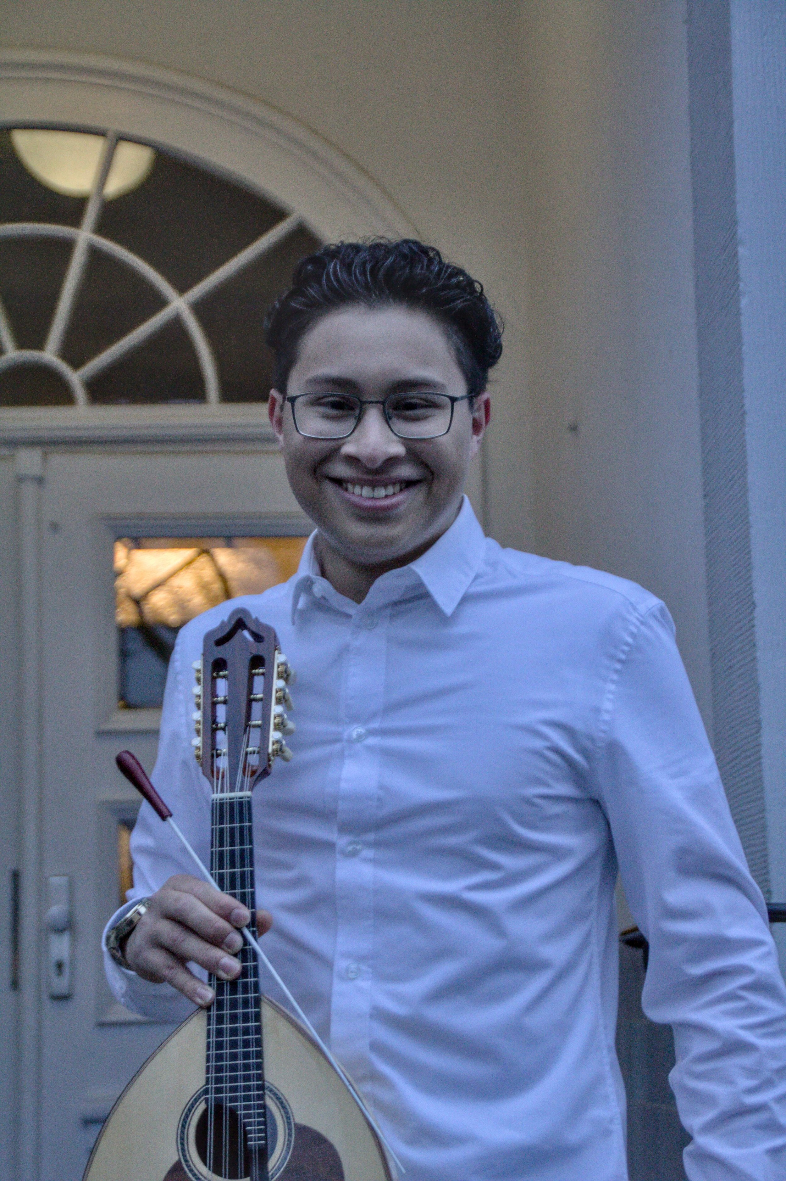 Ein junger Mann trägt eine weiße Hemd und hält eine Geige, er lächelt vor einem Haus mit einer Tür und einem halbkreisförmigen Fenster im Hintergrund.