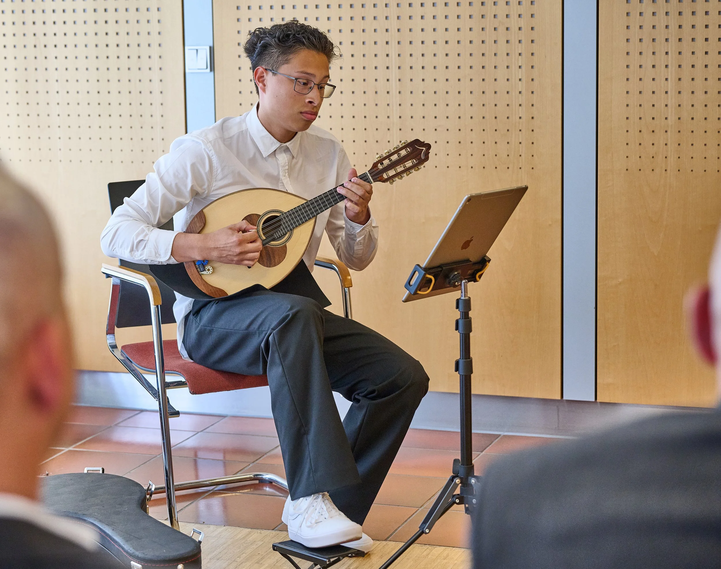 Junger Mann sitzt auf einem Stuhl und spielt Gitarre vor einer Gruppe. Er schaut auf ein Tablet auf einem Ständer. Der Raum hat Holzverkleidungen.