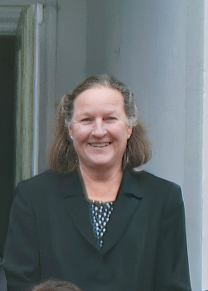 Ein lächelnder Mann mit braunen, schulterlangen Haaren in schwarzer Jacke vor einer grauen Wand.