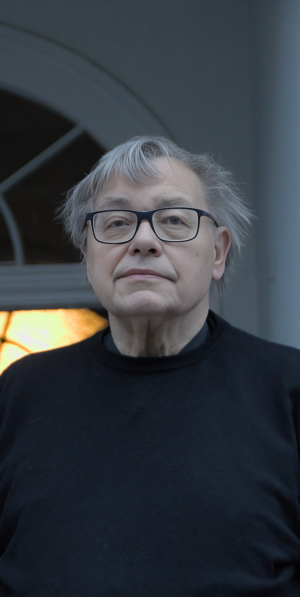 Ein älterer Mann mit grauhaarigem, zerzaustem Haar, trägt schwarze Brille und schwarzen Pullover, steht vor einem dunklen Hintergrund.
