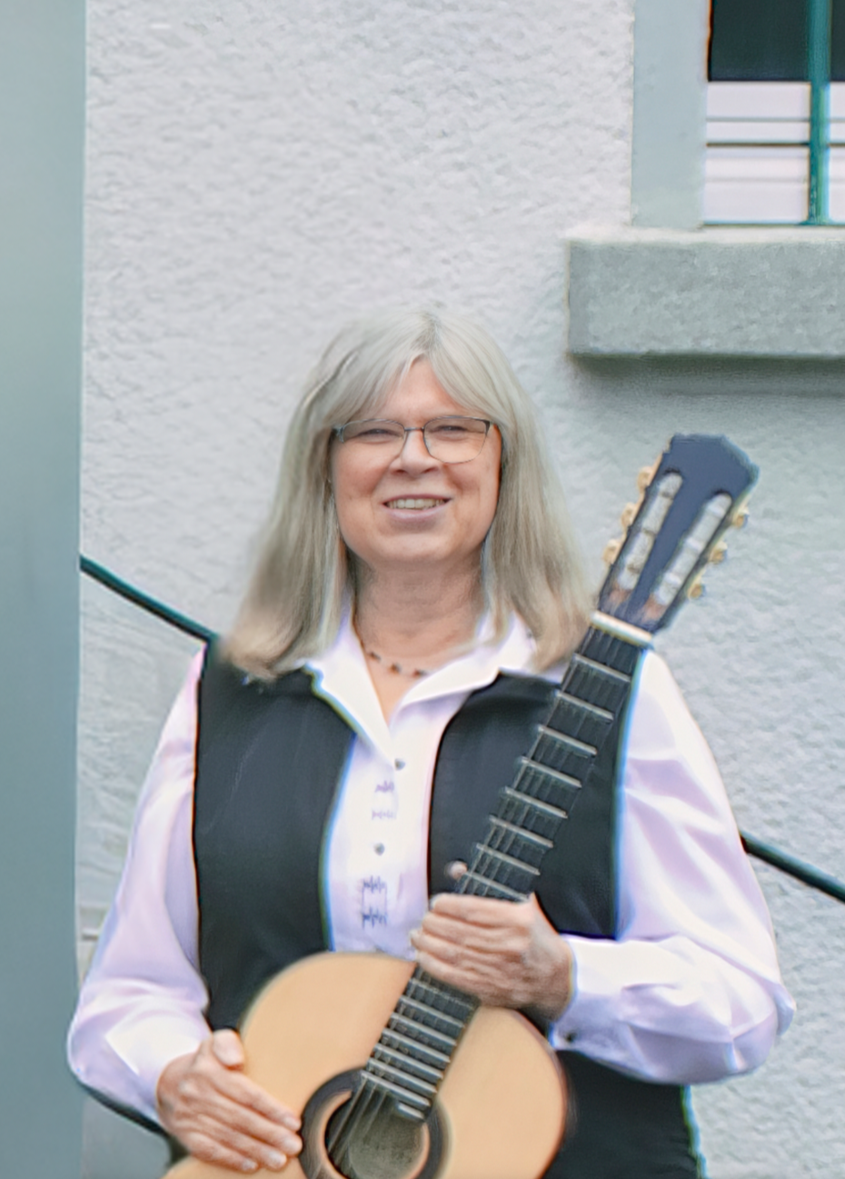 Eine ältere Frau mit langen grauen Haaren und Brille, die ein Akustik-Gitarre in der Hand hält, vor einer weißen Wand mit Balkon. Sie lächelt in die Kamera.