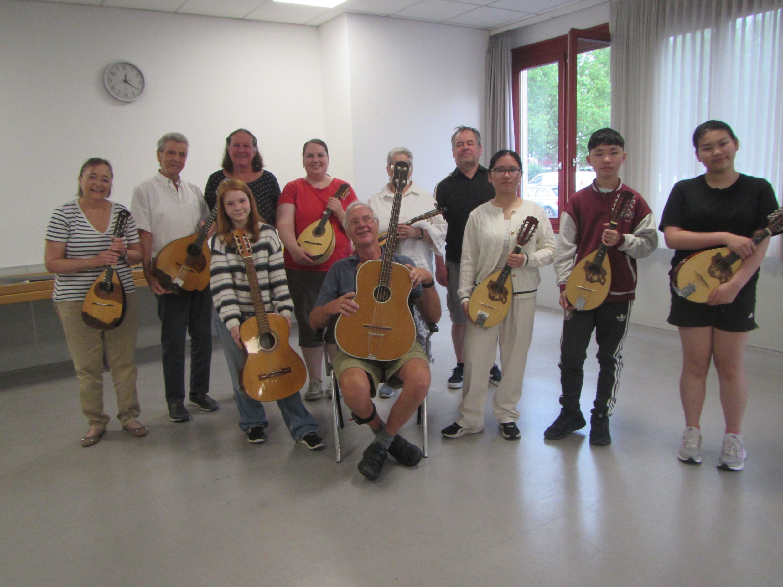 Gruppe von Menschen mit Gitarren in einem Raum mit weißen Wänden, großen Fenstern und einem Wanduhr.