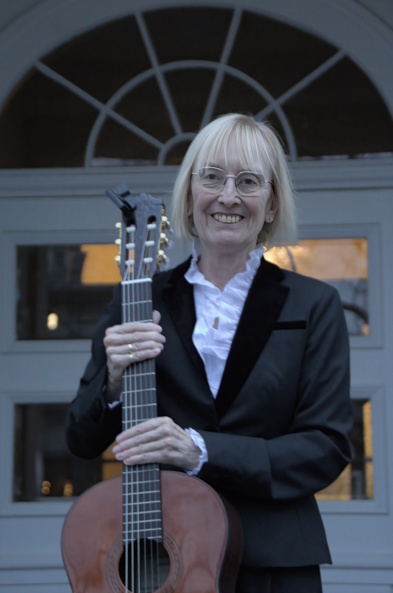 Eine lächelnde ältere Frau mit Kurzhaarfrisur und Brille, die eine Gitarre hält, steht vor einer Tür mit einem halbkreisförmigen Fenster. Sie trägt einen schwarzen Blazer und ein weißes Hemd.