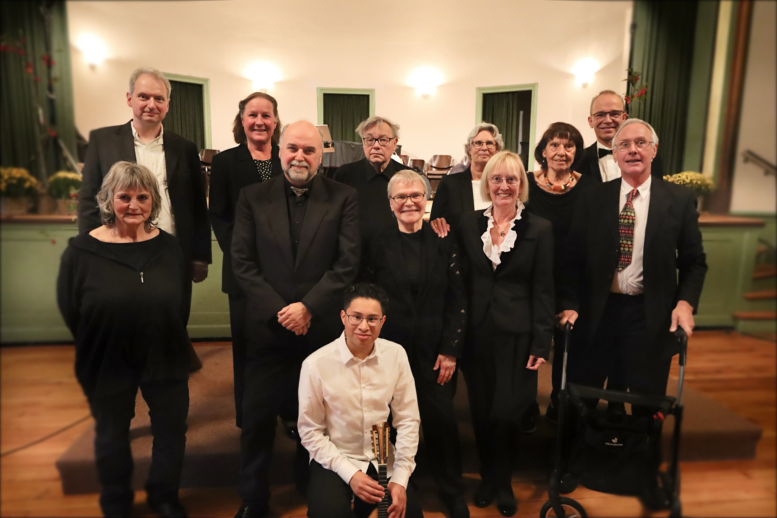Gruppe von 12 Menschen bei einer Veranstaltung, einige in formeller Kleidung, in einem Saal mit grünen Wänden und Holzfußboden.