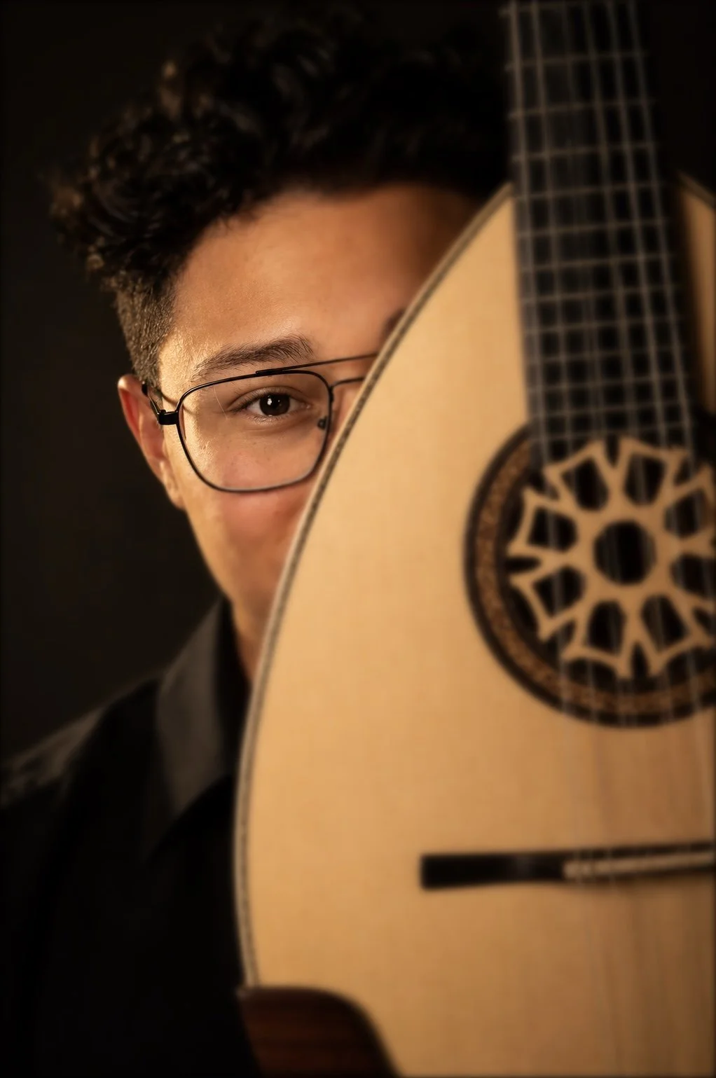 Ein junger Mann mit Brille hält eine Akustikgitarre, sein Gesicht halb hinter der Gitarre verborgen, gegen einen dunklen Hintergrund.