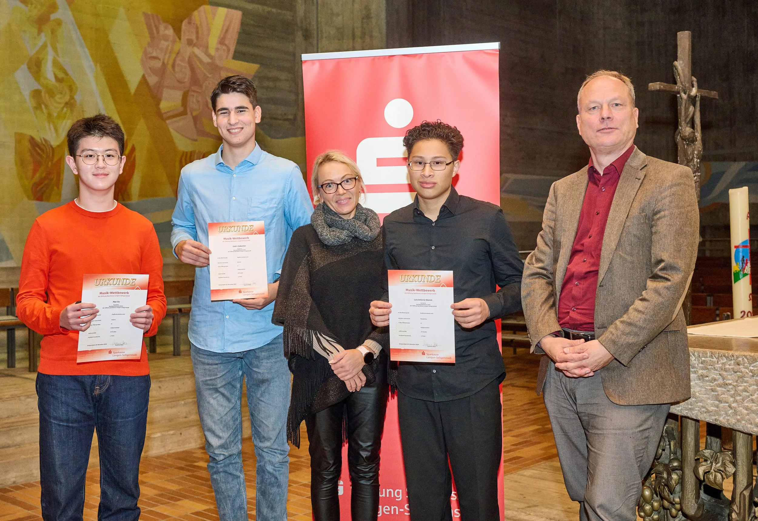Vier Personen stehen in einer Halle, drei von ihnen halten Urkunden. Es sieht aus, als ob sie bei einer Preisverleihung oder einem Wettbewerb teilnehmen. Hinter ihnen ist ein rotes Banner mit einem weißen Symbol, wahrscheinlich eine Organisation oder Firma. Im Hintergrund sind Wandbilder und eine Statue sichtbar.