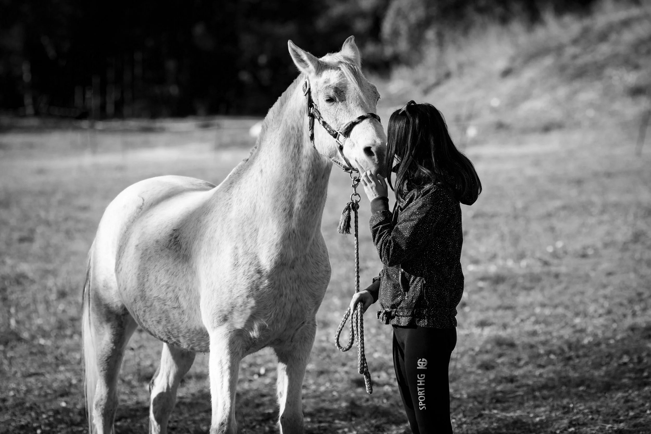 Une fille avec des cheveux noirs courts, portant un jean de sport, caresse un cheval blanc dans un champ en plein air.