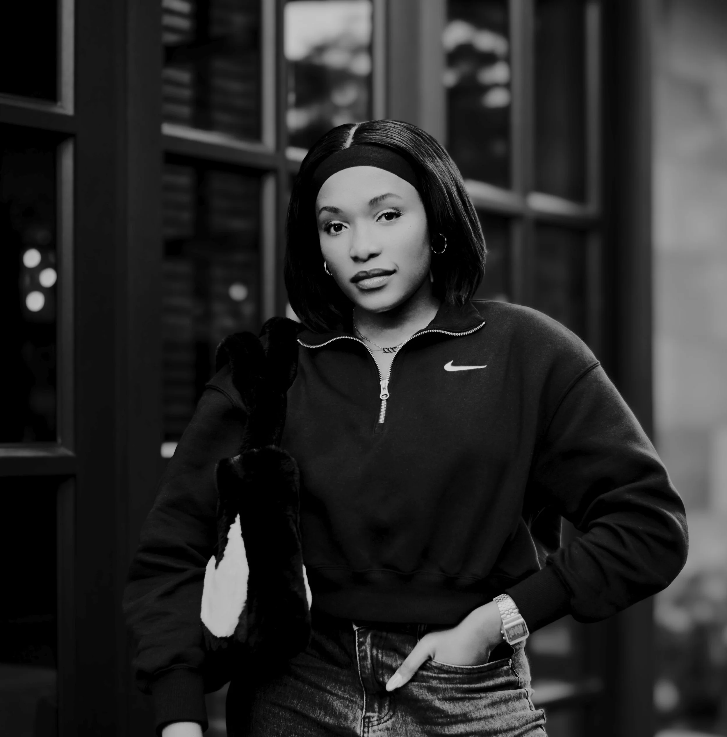 Black and white photo of a young woman with shoulder-length hair, wearing a Nike zip-up sweatshirt, earrings, and a watch, standing with one hand in her pocket in front of a bookshelf.